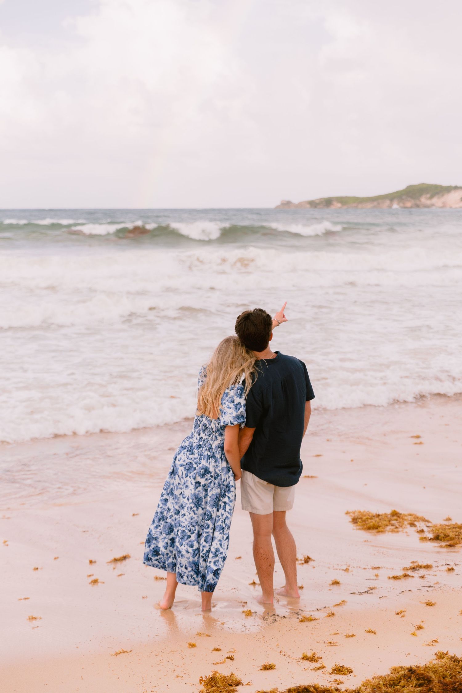 A couple walks along a sandy beach with crashing waves and seaweed on a cloudy day.