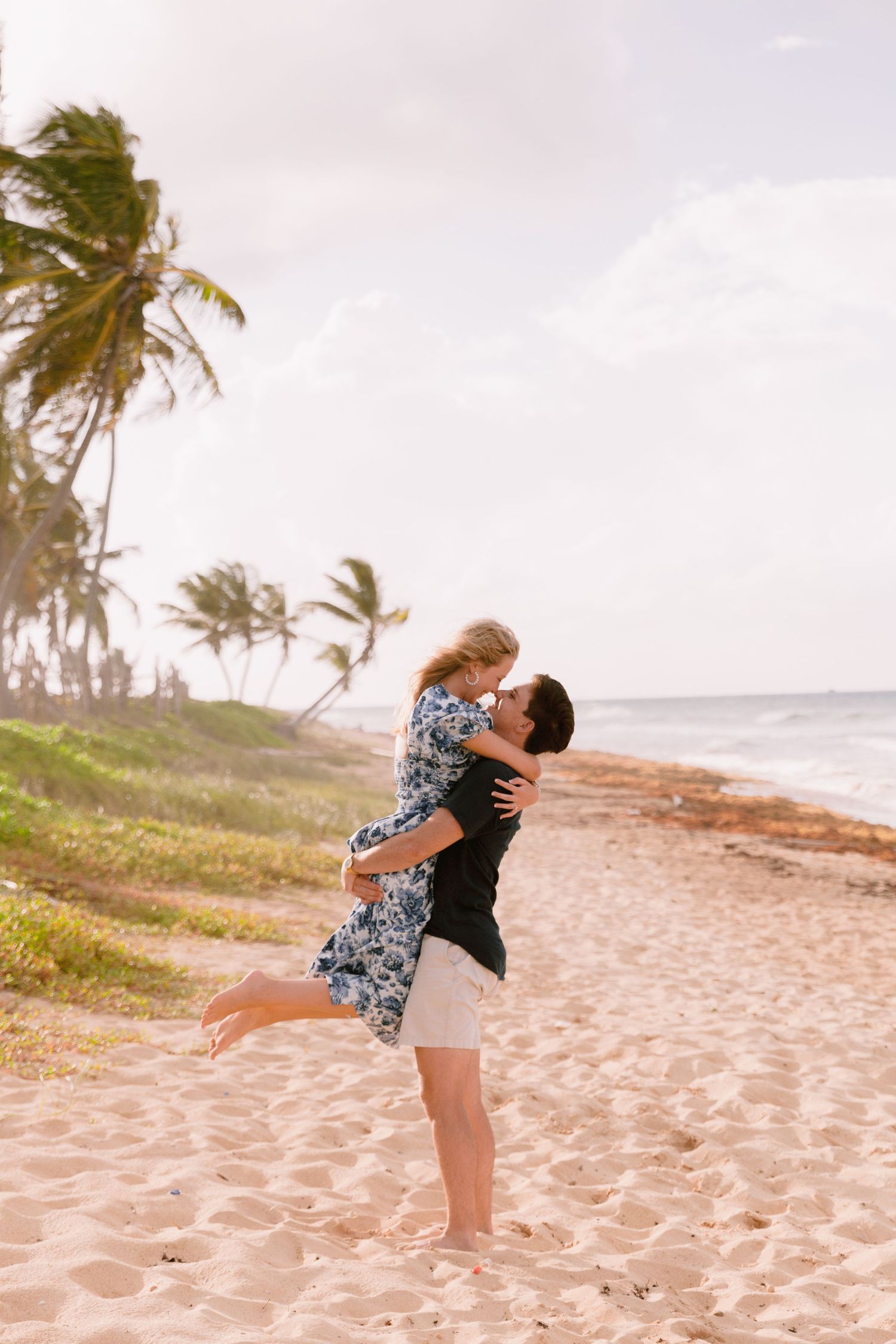 Two people embrace lovingly on a tropical beach with palm trees swaying in the wind at sunset.