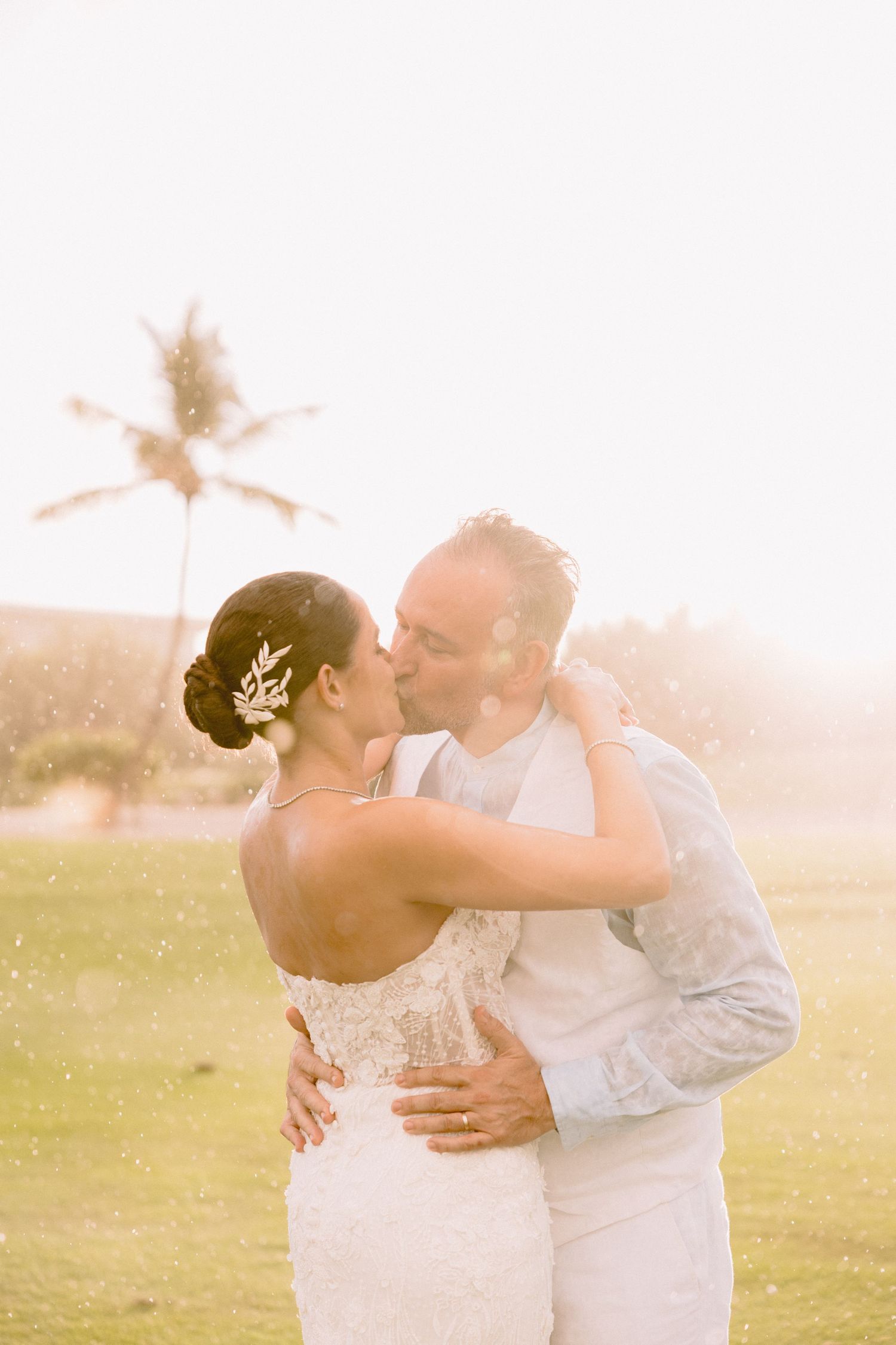 A romantic sunset wedding photoshoot shows a couple embracing in a sun-drenched field with palm trees in the background.