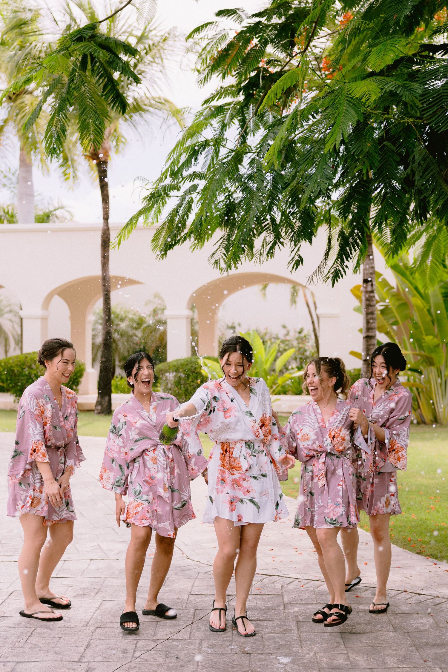 A group in matching floral robes walks and laughs together on a stone path under palm trees at a tropical resort.