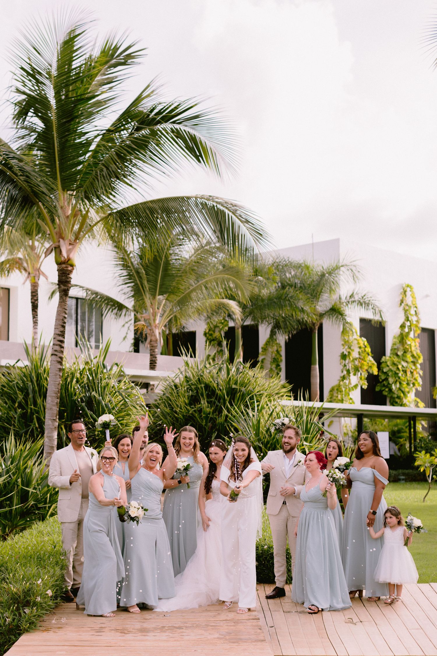 Wedding party in dusty blue and beige attire posing on wooden walkway surrounded by tropical palm trees.