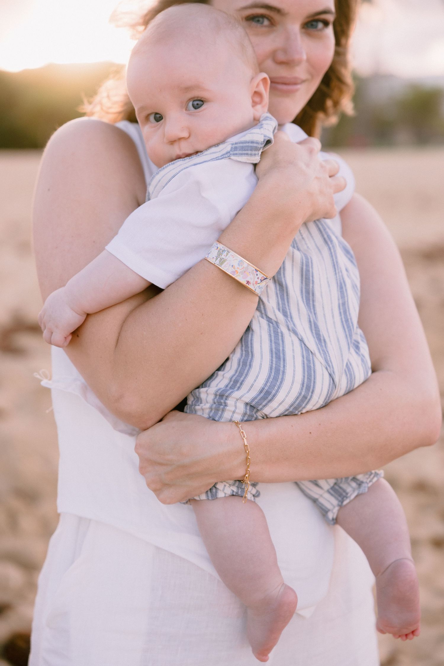 A mother holds her baby on a sunny beach at golden hour, both wearing casual summer outfits.