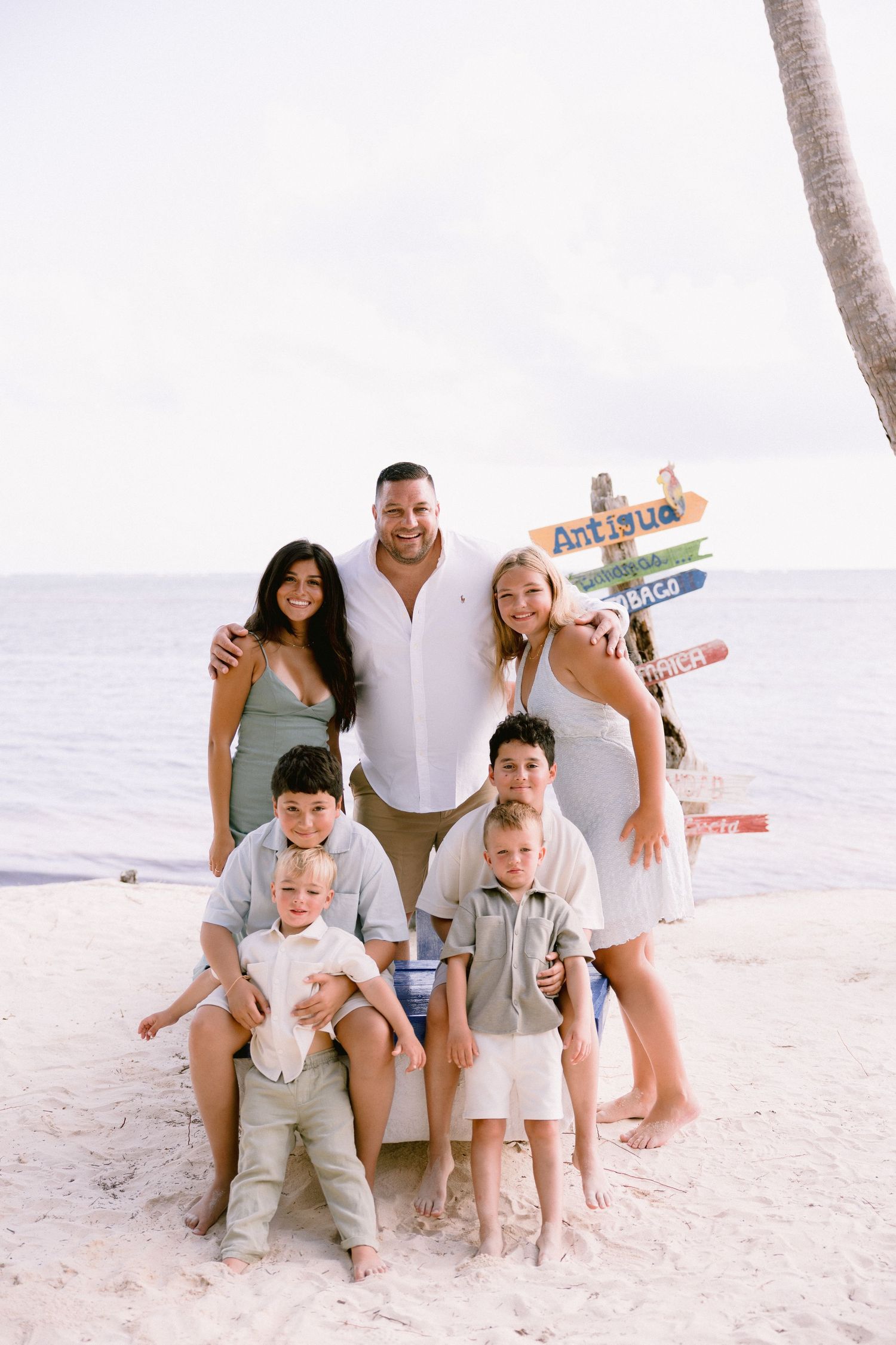 Family of six posing on a sandy beach near a colorful directional sign with ocean views in the background.