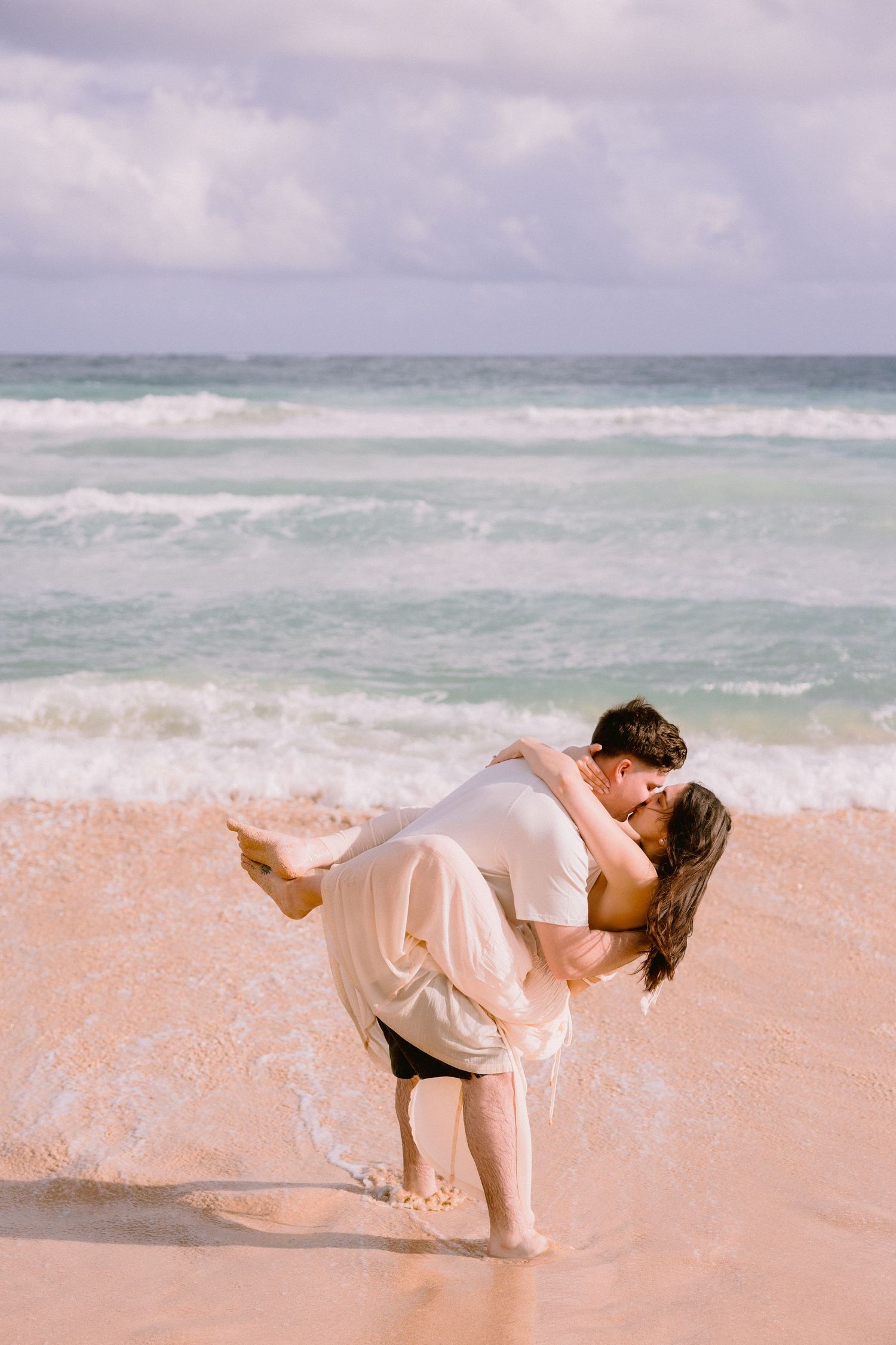 Romantic couple sharing a kiss on a tropical beach, man dipping woman near ocean waves on sandy shore.