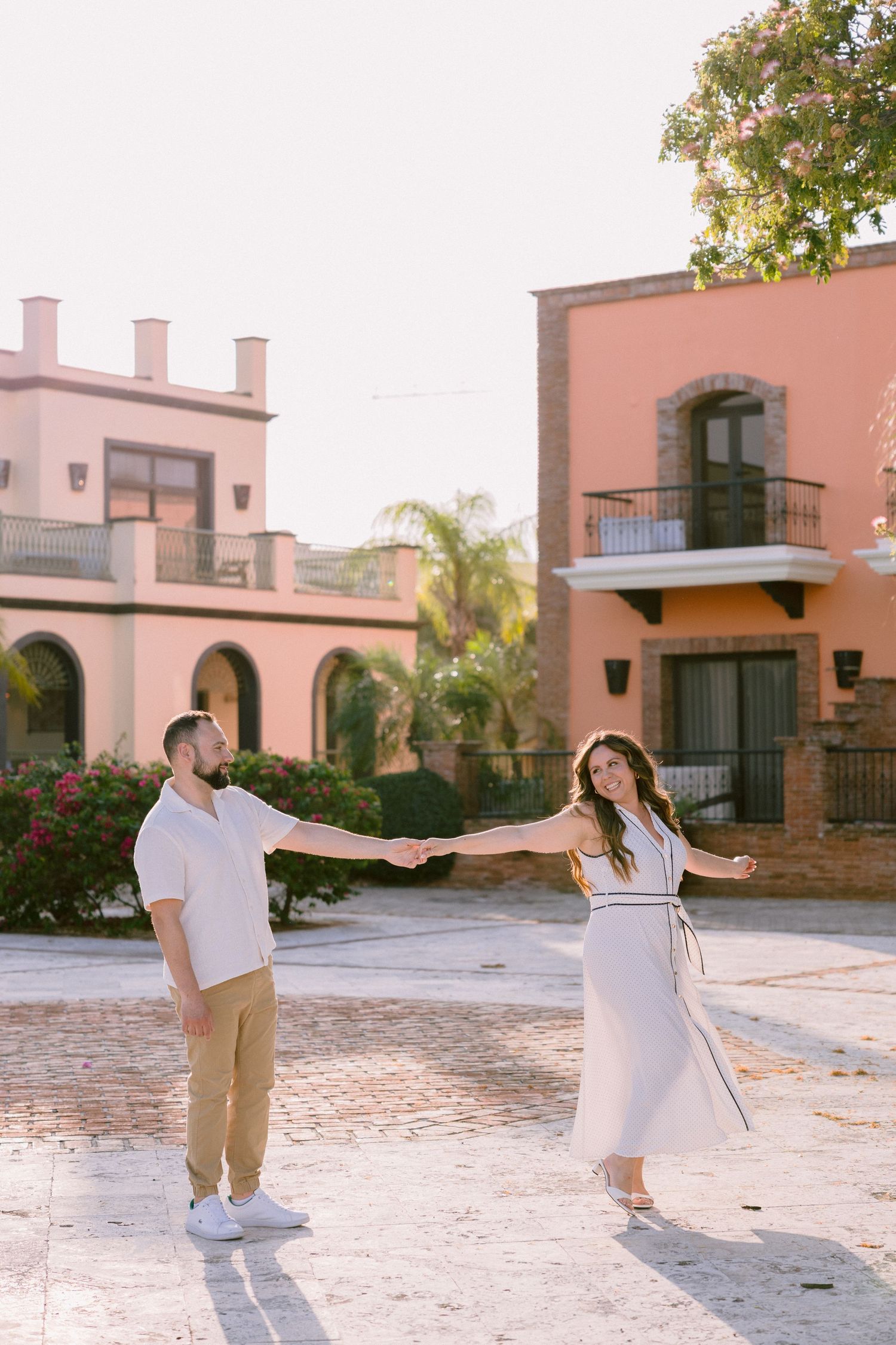 Couple holding hands and dancing in a sunlit Mexican courtyard, woman in white dress, man in white shirt and tan pants.