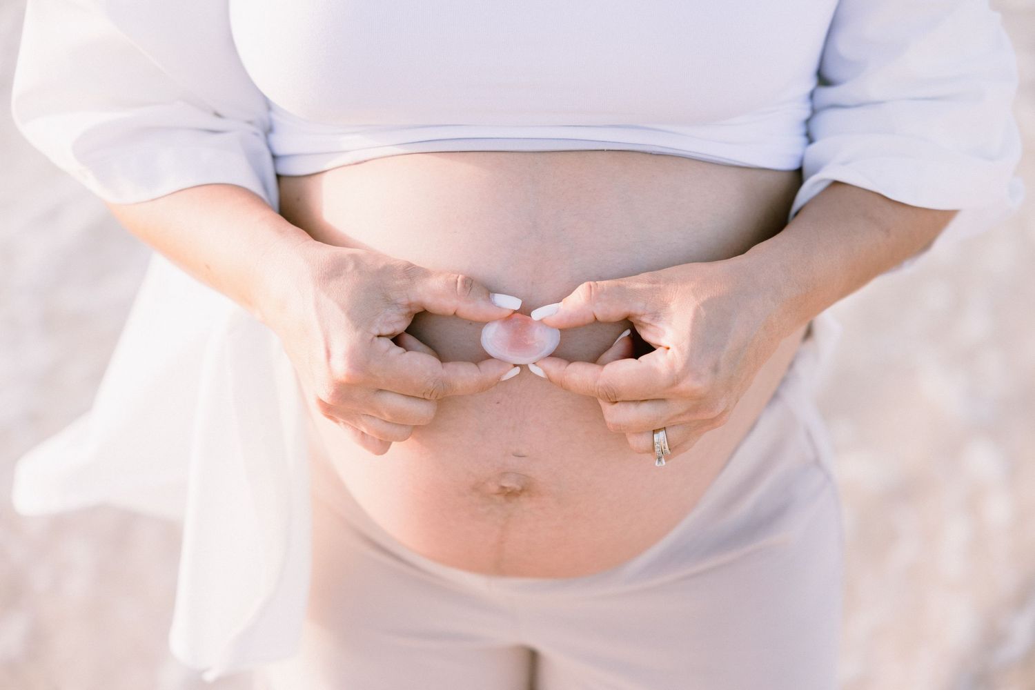 Pregnant woman holding baby shoes over her bare belly, wearing a white top and beige pants outdoors.