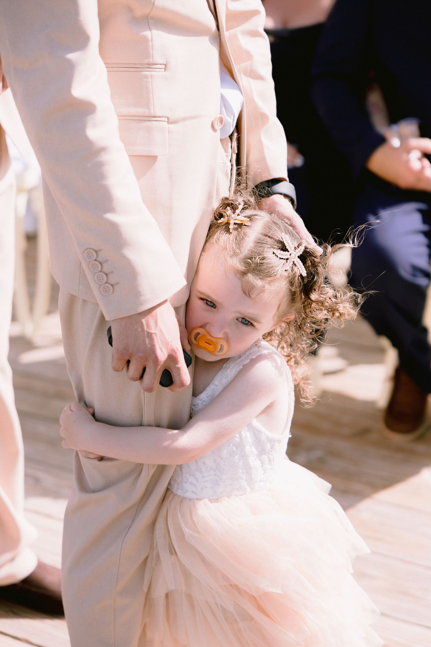 Adorable flower girl in blush tulle dress hugging a man in beige suit at a wedding, with a pacifier.