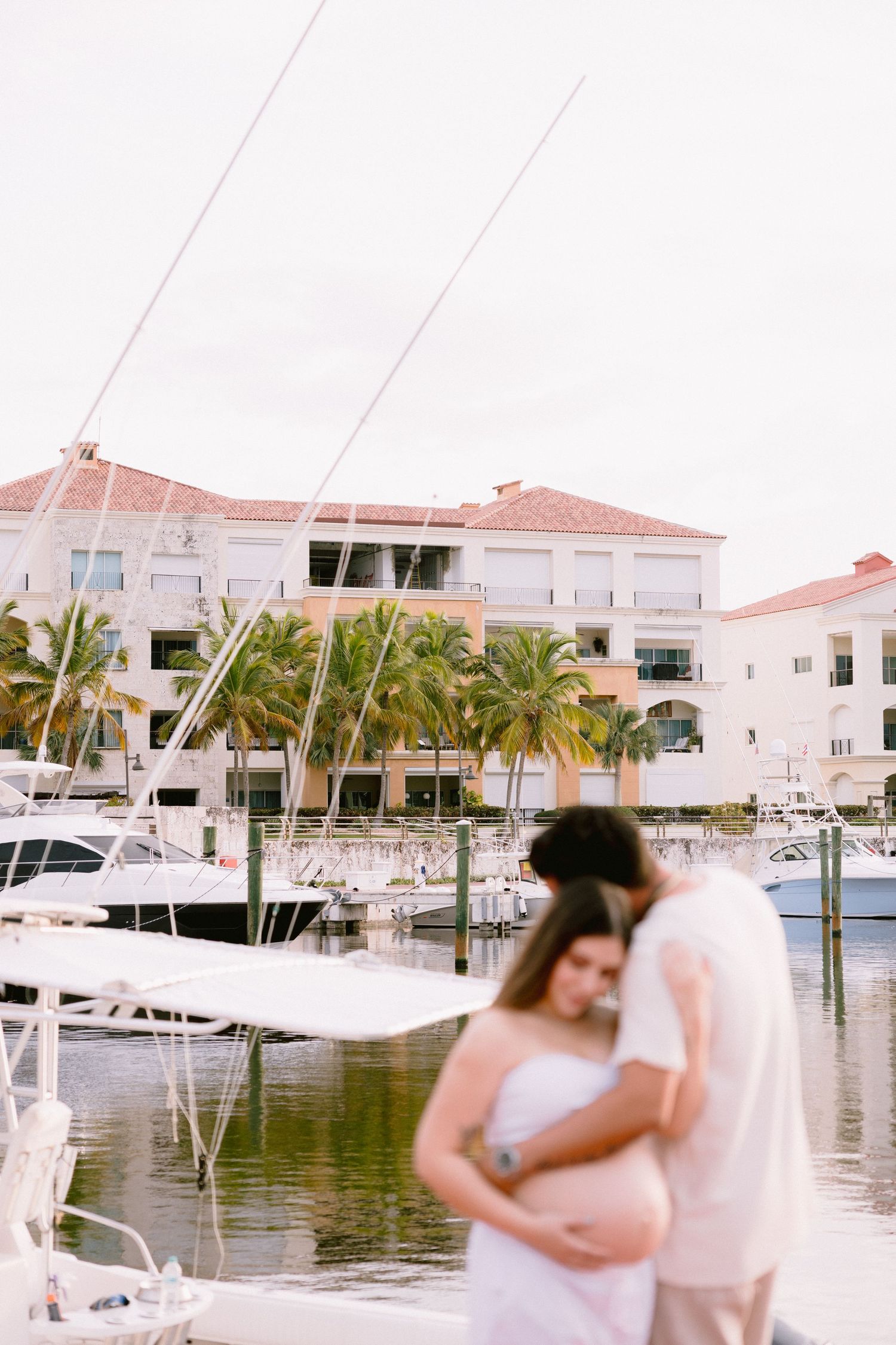 A scenic marina with white boats docked along the waterfront near palm trees and beige waterfront buildings.