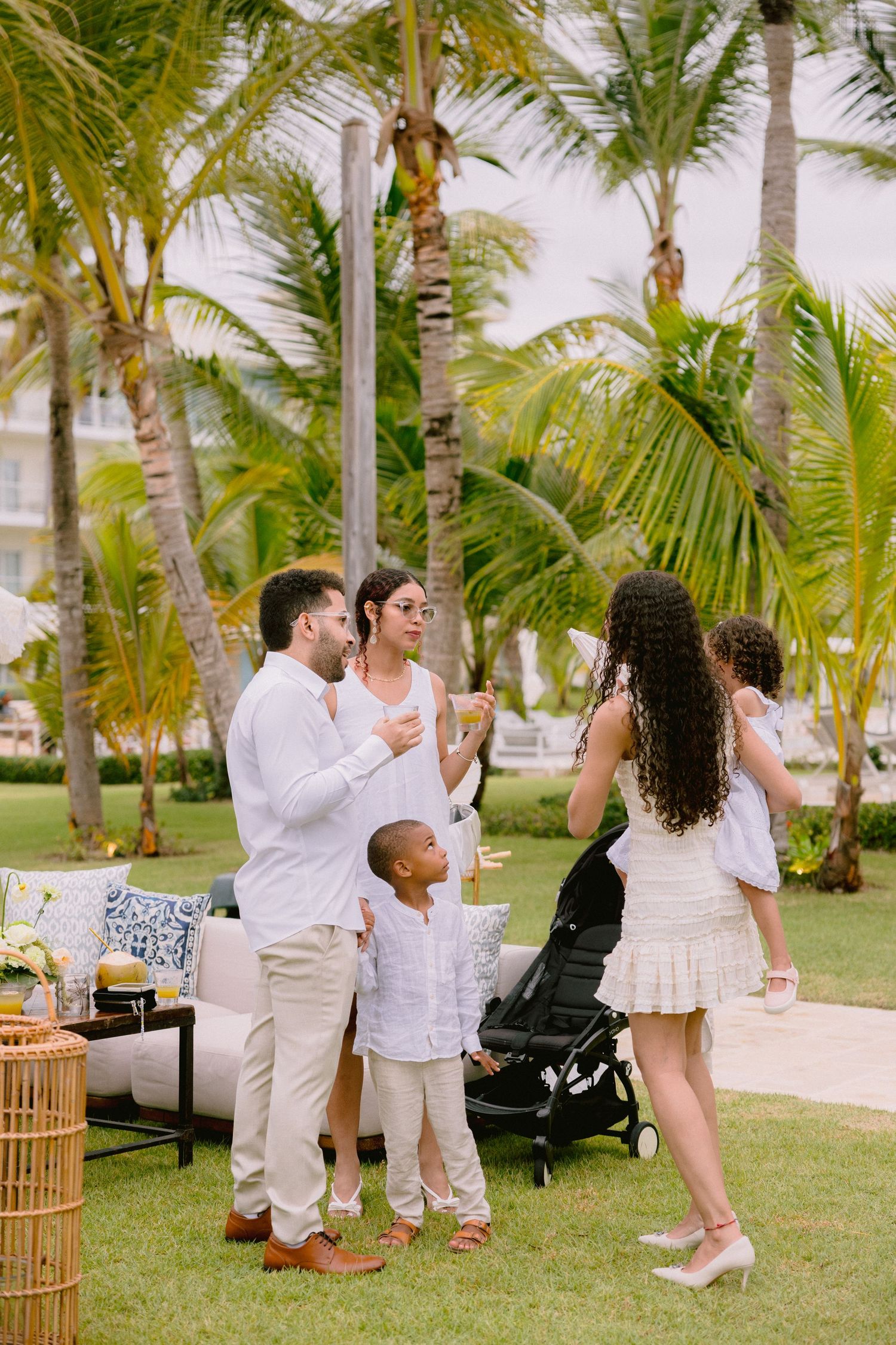 Family in white and neutral outfits gathers on a tropical resort lawn with palm trees, a stroller, and lounge seating nearby.