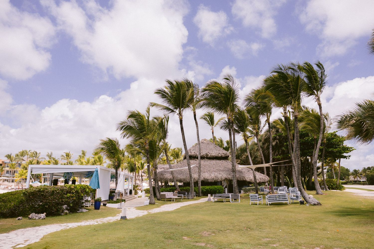 Tropical resort grounds with tall palm trees, a thatched-roof palapa, lounge chairs and a stone pathway under a bright blue sky.