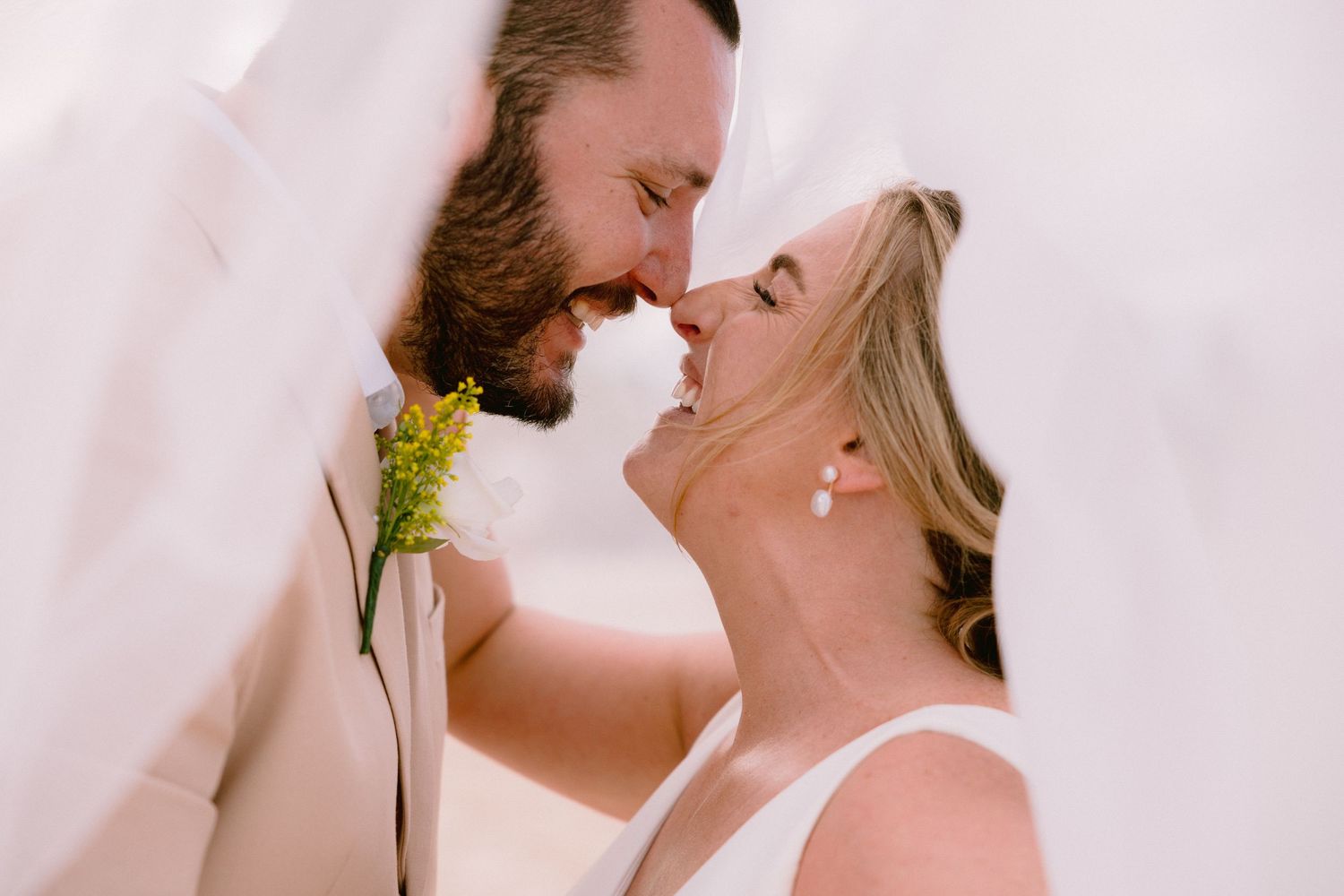 A bride and groom share a romantic moment under a white veil, nearly kissing, with a yellow boutonniere visible.