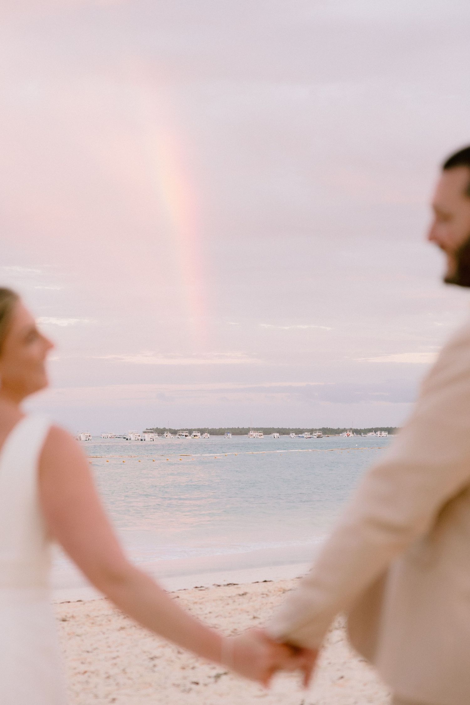 Couple holding hands on beach with rainbow over ocean at sunset wedding ceremony.