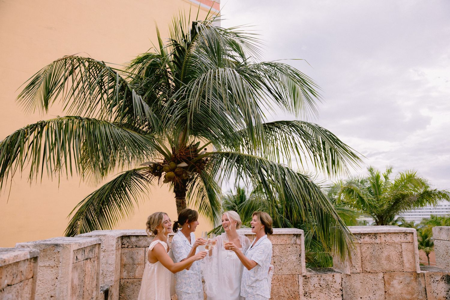 Bridesmaids in white dresses toasting near a stone wall with tropical palm trees at a destination wedding.