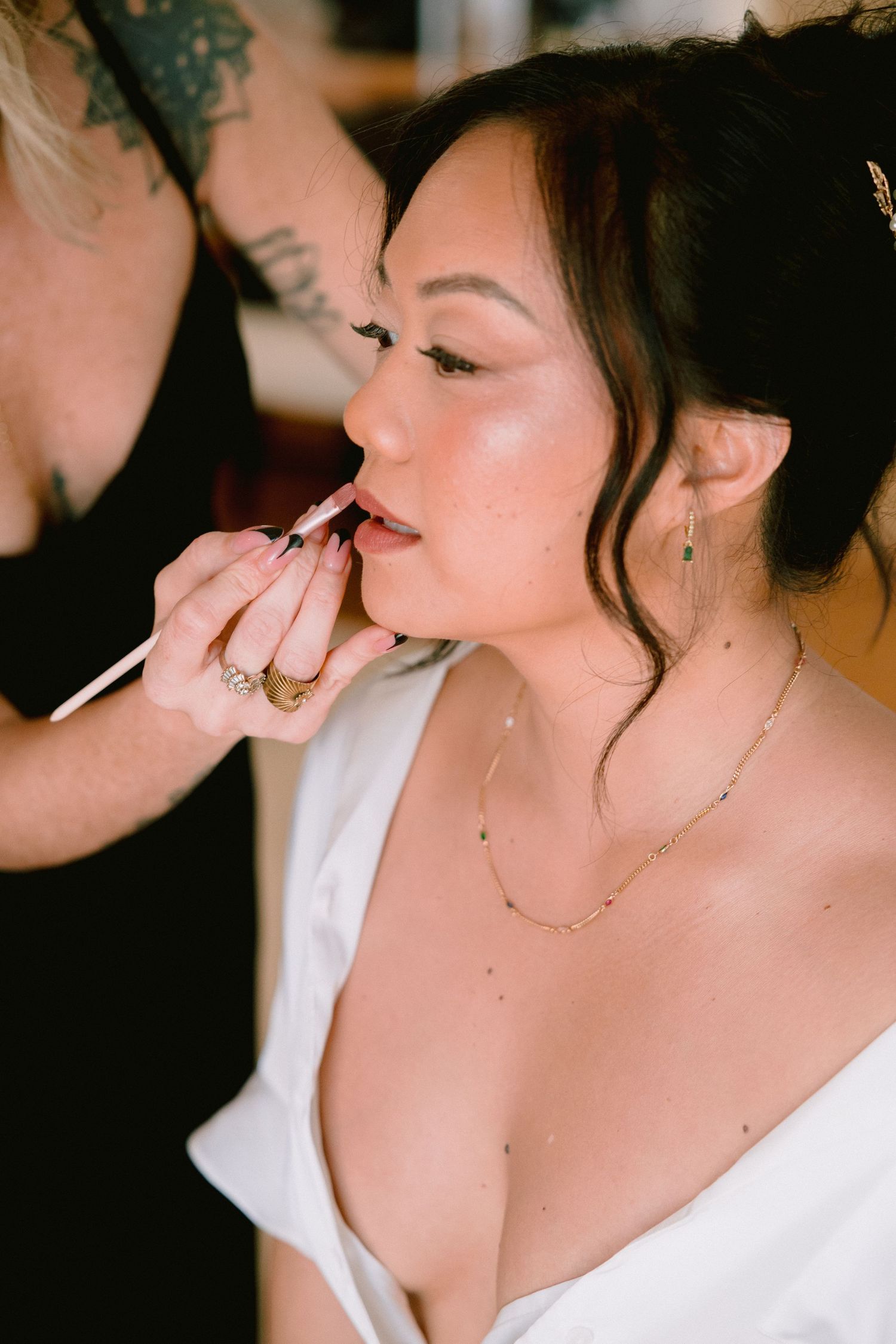 A makeup artist applying lipstick to a bride in a white dress, wearing gold jewelry, with an elegant updo hairstyle.