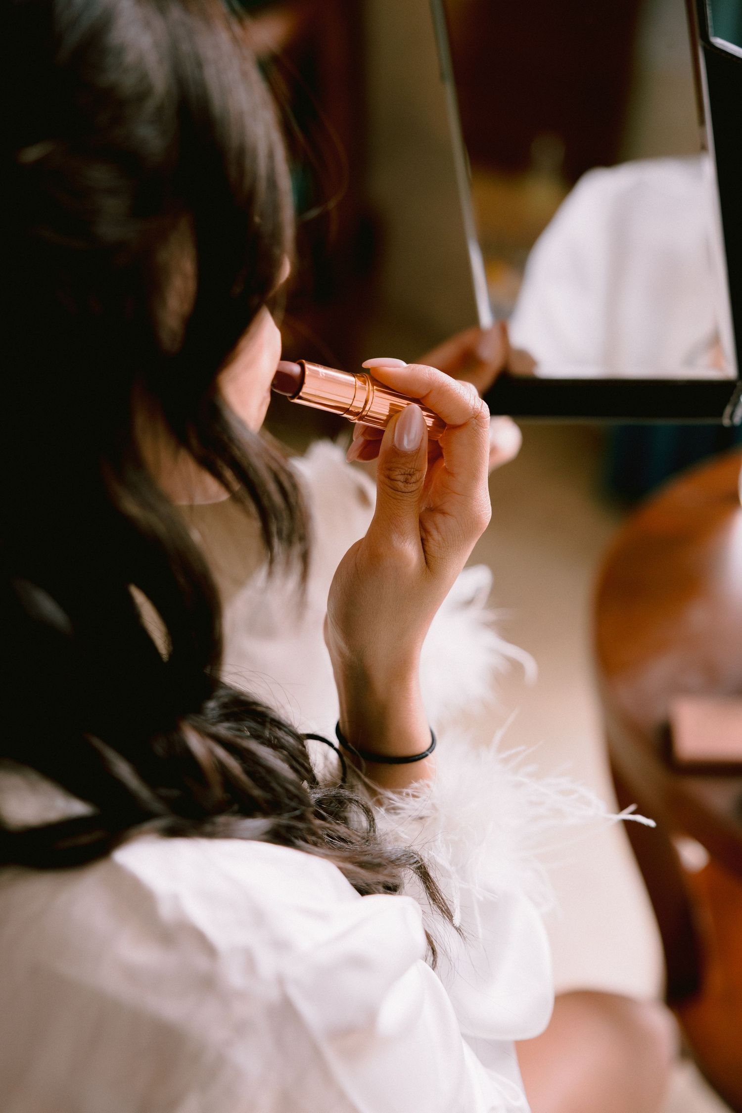 Woman in white robe applying nude lipstick while looking in mirror during bridal getting-ready routine.