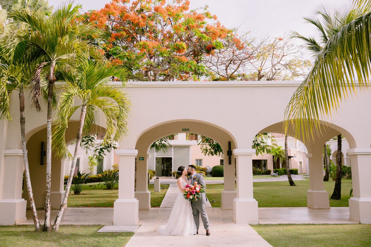 White stone archways line a tropical garden path with palm trees and vibrant orange flowering plants.