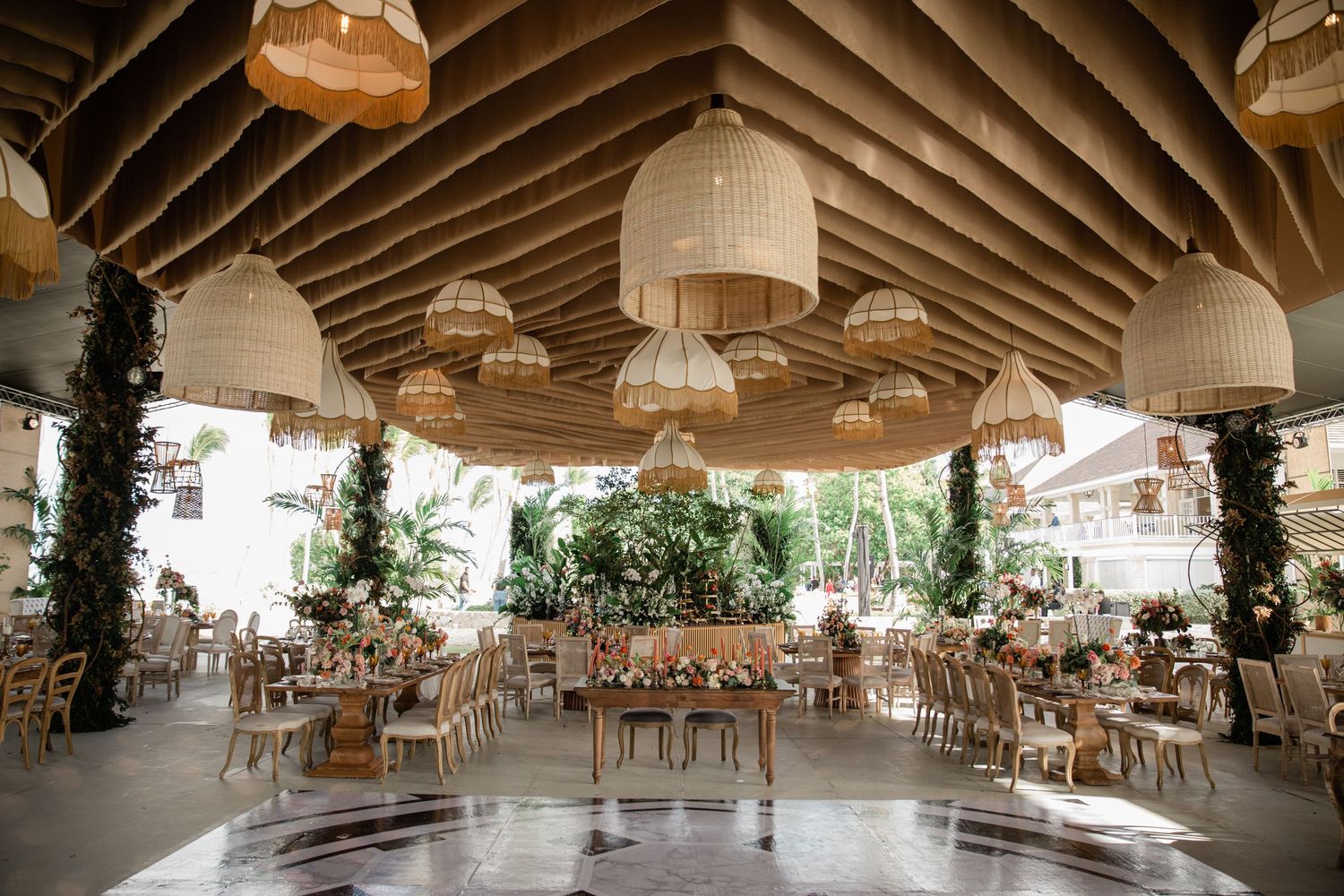 An elegant wedding reception venue with wooden tables, hanging pendant lights, and greenery-wrapped pillars under a vaulted ceiling.