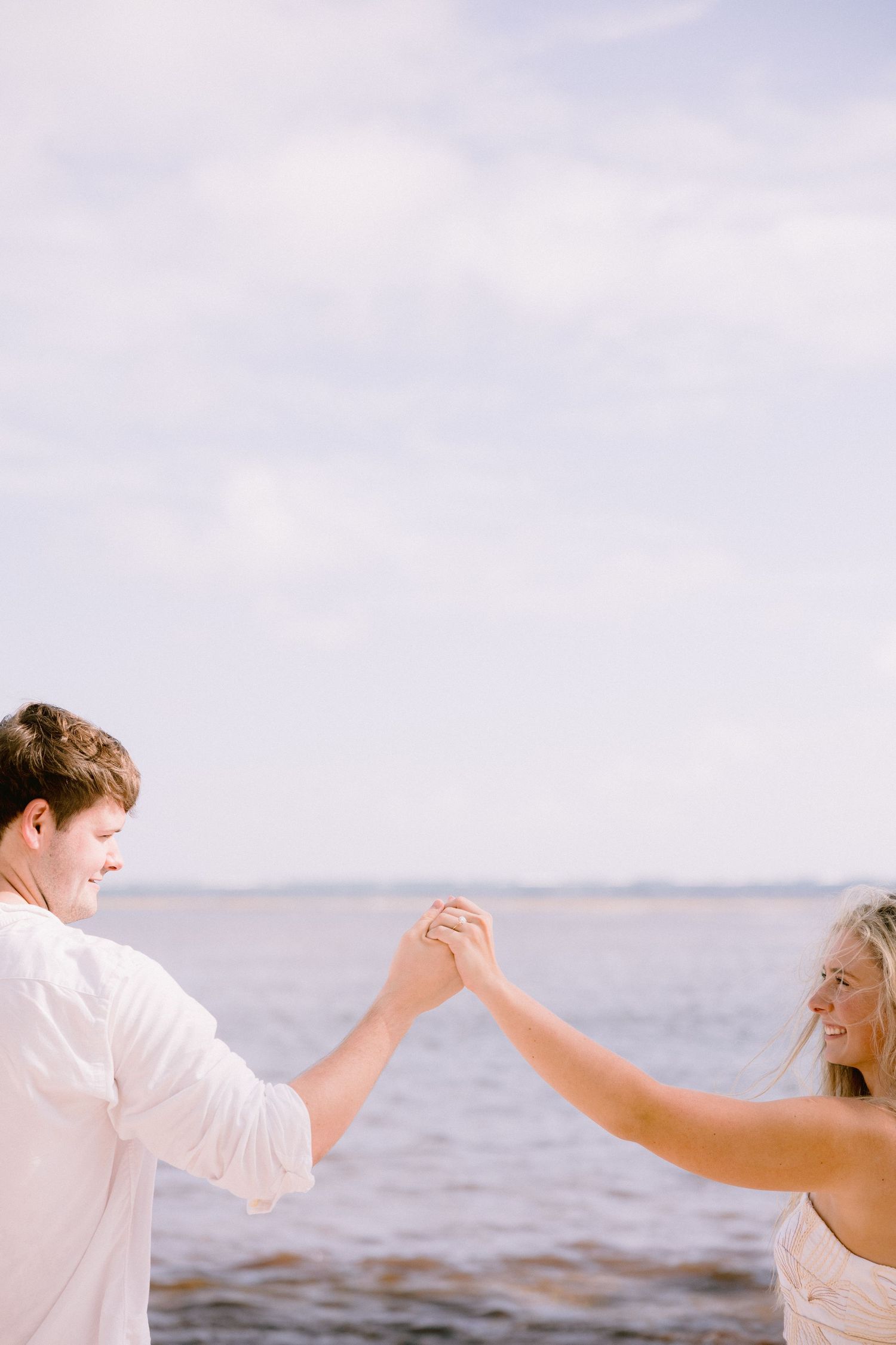 A couple in white clothing dances playfully together on the beach with the ocean in the background.
