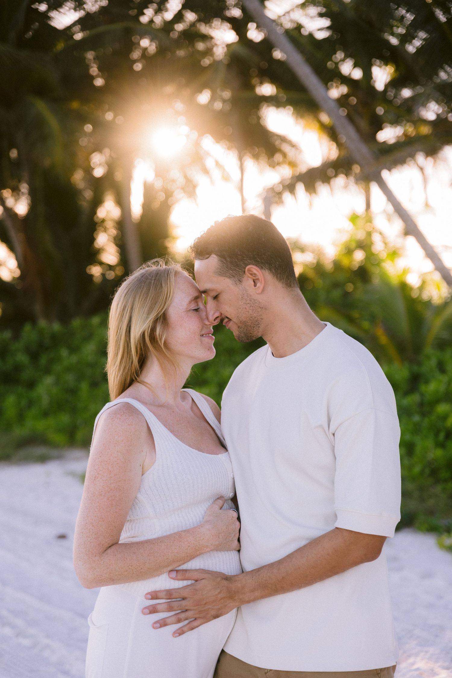 Couple in white outfits sharing a romantic moment on a beach at sunset with tropical palm trees.