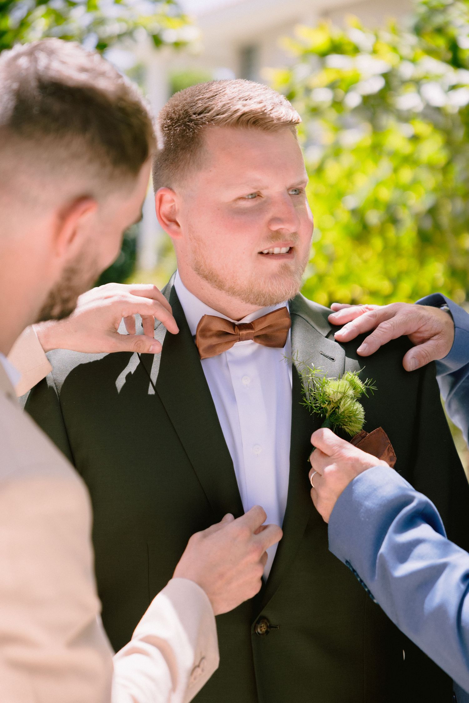 Groomsmen helping groom adjust his dark suit, brown bow tie, and boutonniere outdoors before a wedding ceremony.