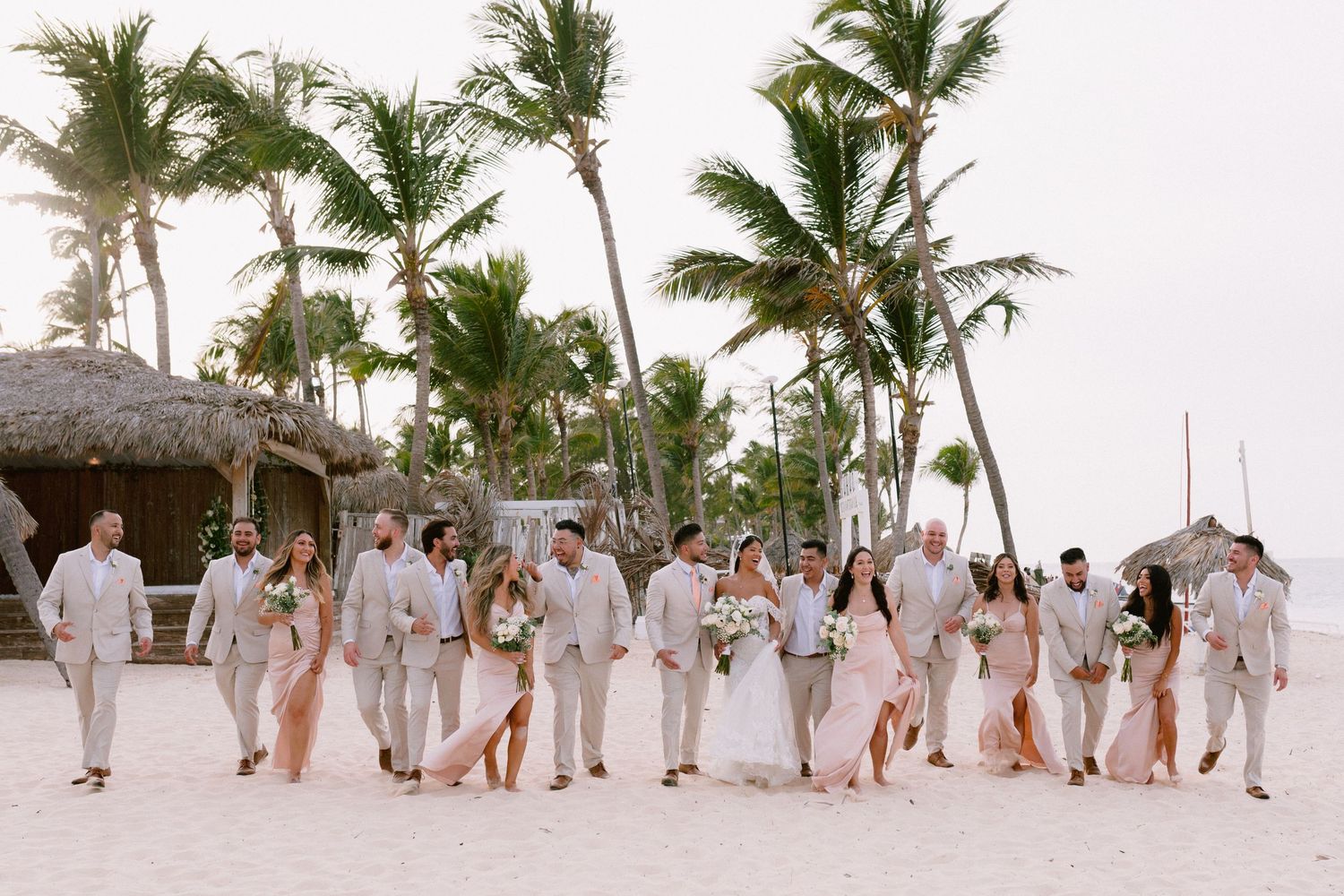 Wedding party walks on tropical beach with palm trees, bride and groom surrounded by groomsmen in tan suits and bridesmaids in blush gowns.