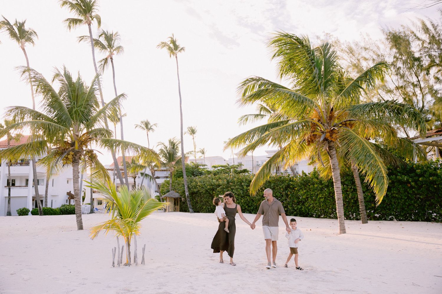 Family of three walking hand-in-hand on a white sandy beach surrounded by tall palm trees at sunset.