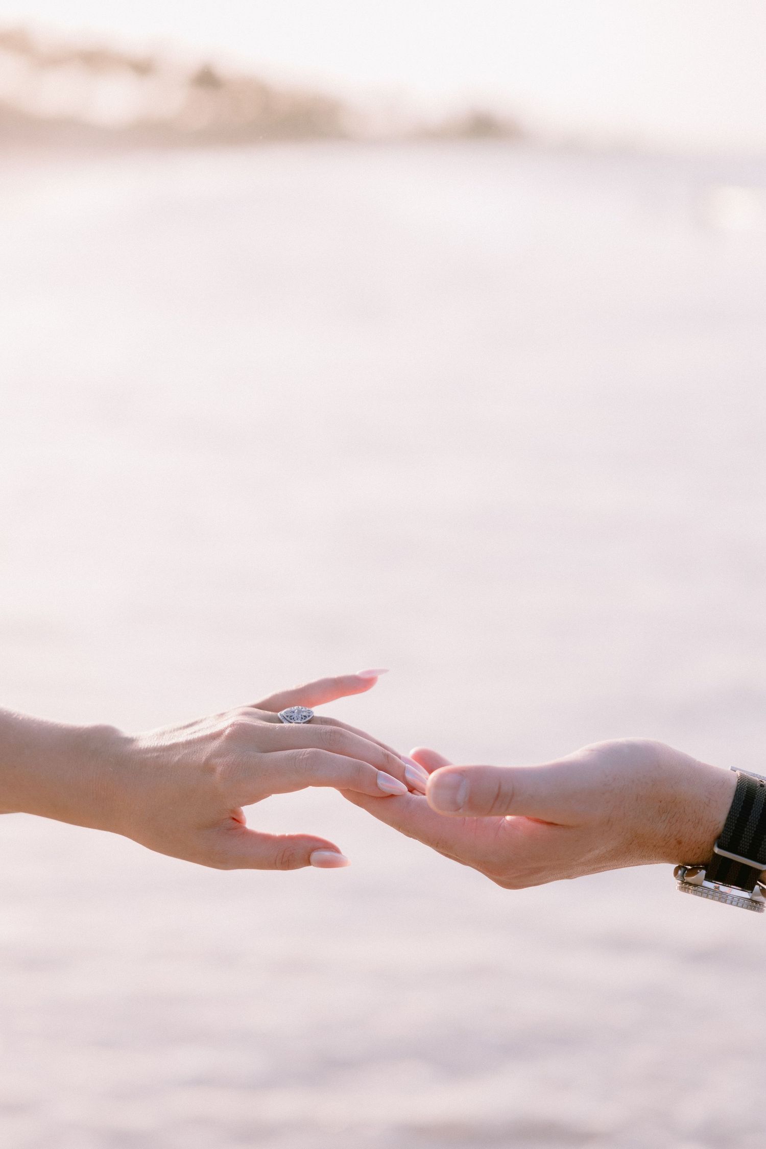 Two hands gently touching near a beach, one adorned with a blue gemstone ring, in soft golden light.