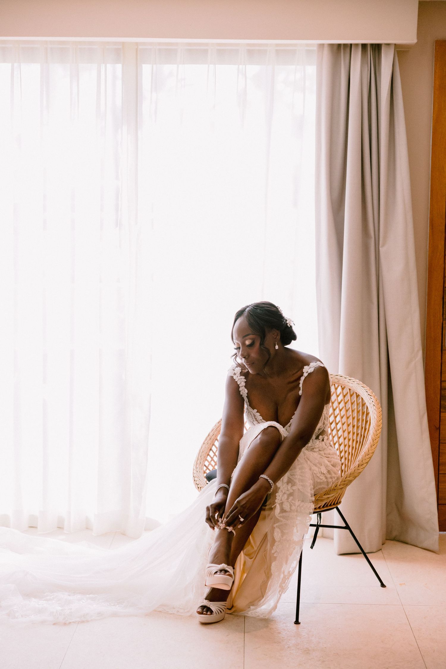 A person in a white dress sits thoughtfully in a woven chair by a sunlit window with sheer curtains.