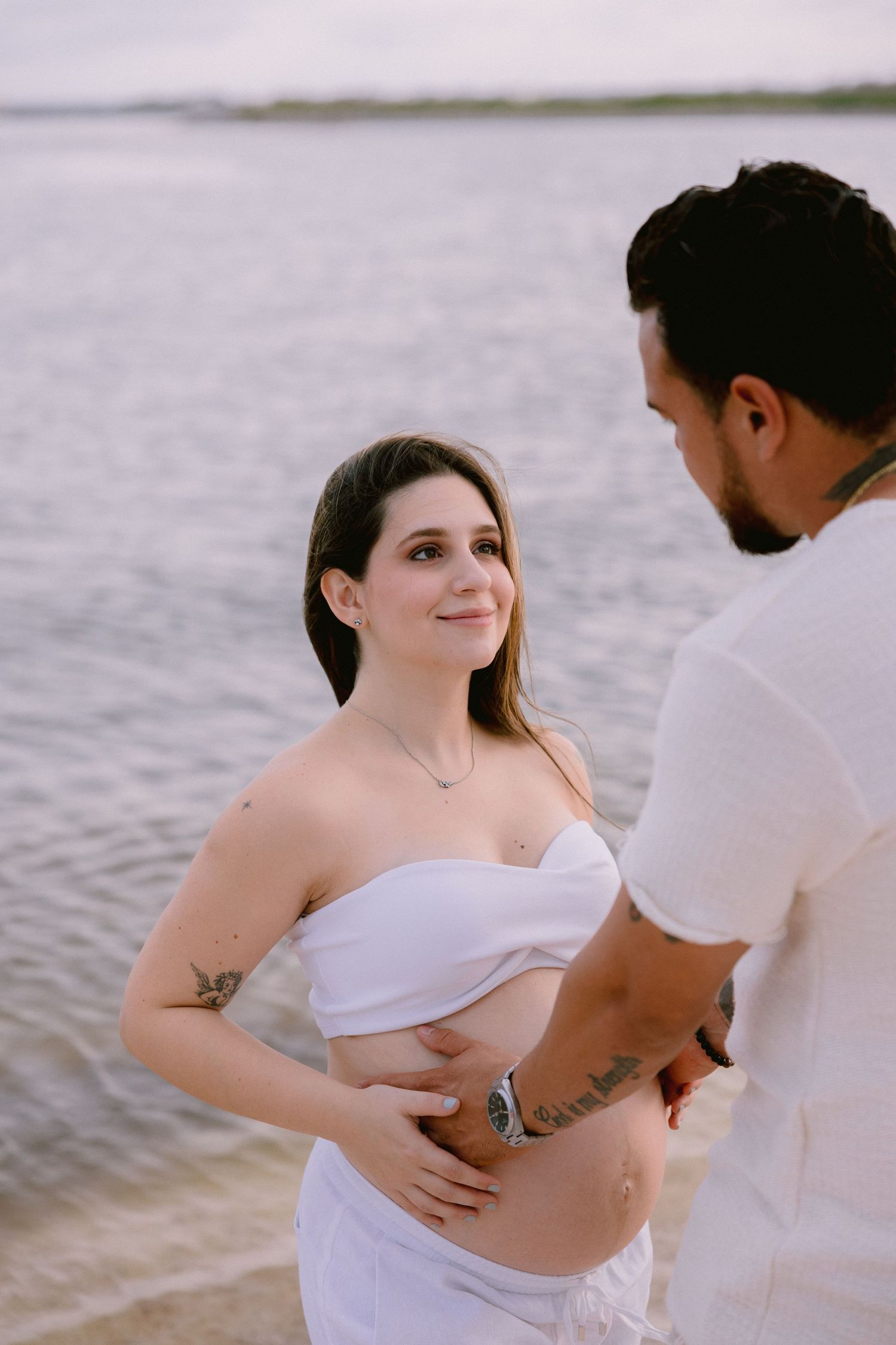 Intimate maternity photoshoot on a beach showing tender moments between an expectant couple in white attire.