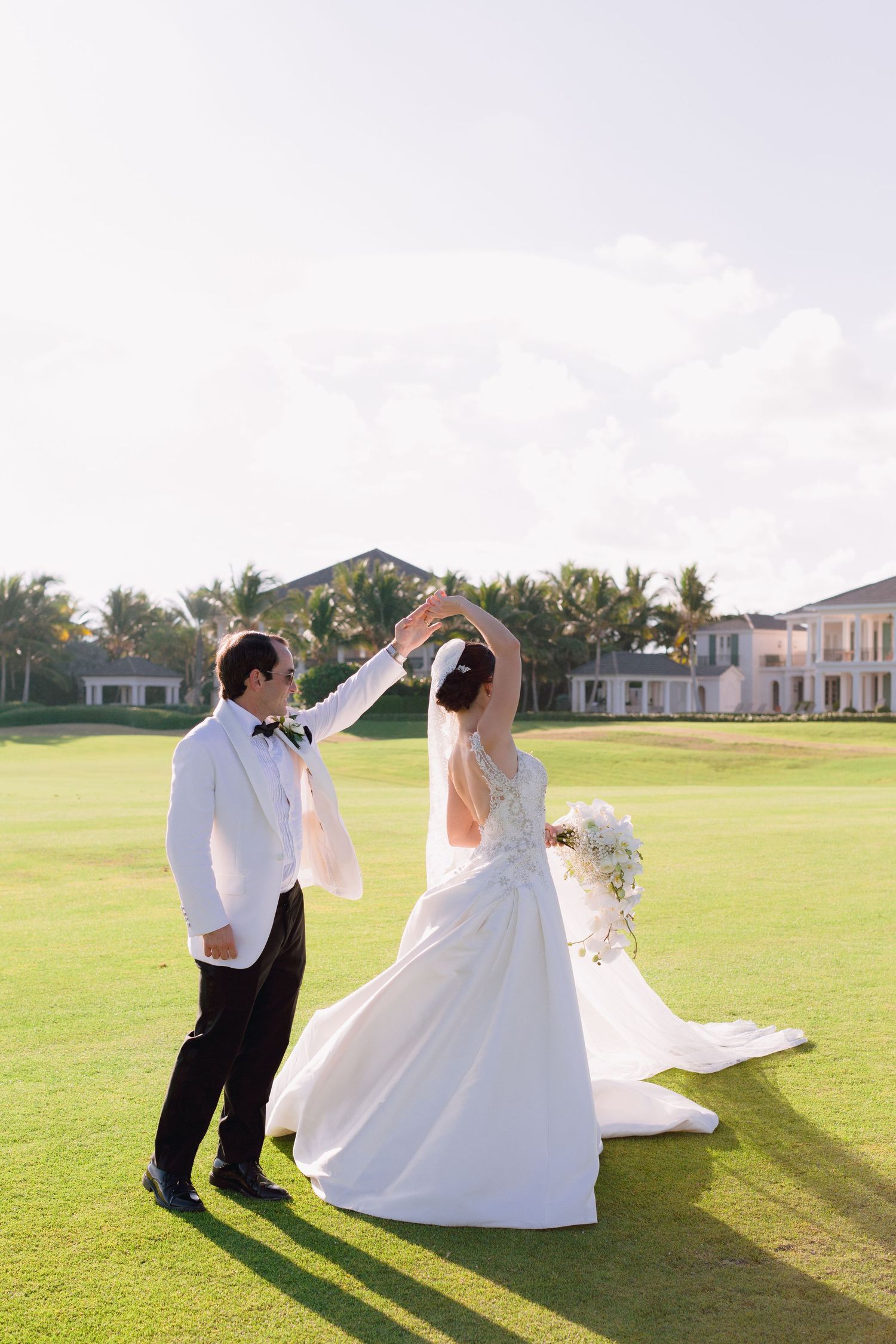 A couple in wedding attire dances and twirls together on a sunny golf course with palm trees in the background.