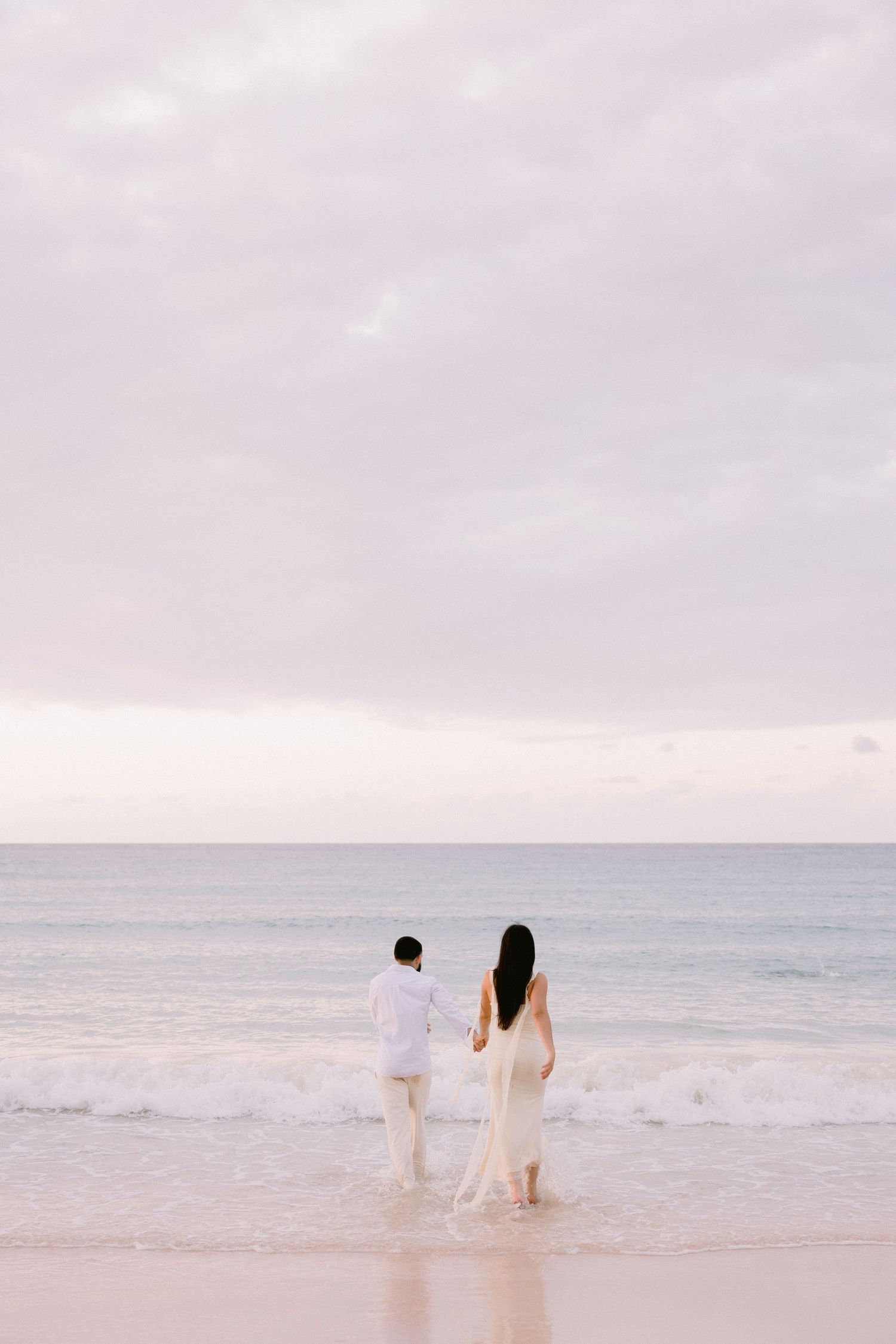 A couple in white attire holds hands while wading in ocean waves on a serene beach at dusk.