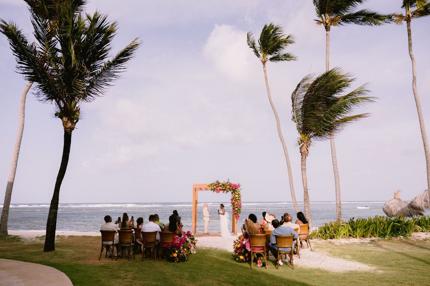 A beachside wedding ceremony setup with palm trees swaying in the wind and guests seated on a grassy lawn.