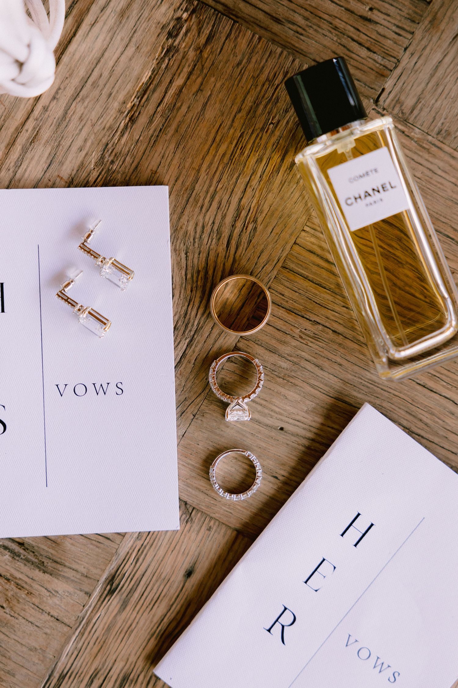 Wedding details flatlay with Chanel perfume, gold earrings, wedding rings and vows cards on wooden surface.