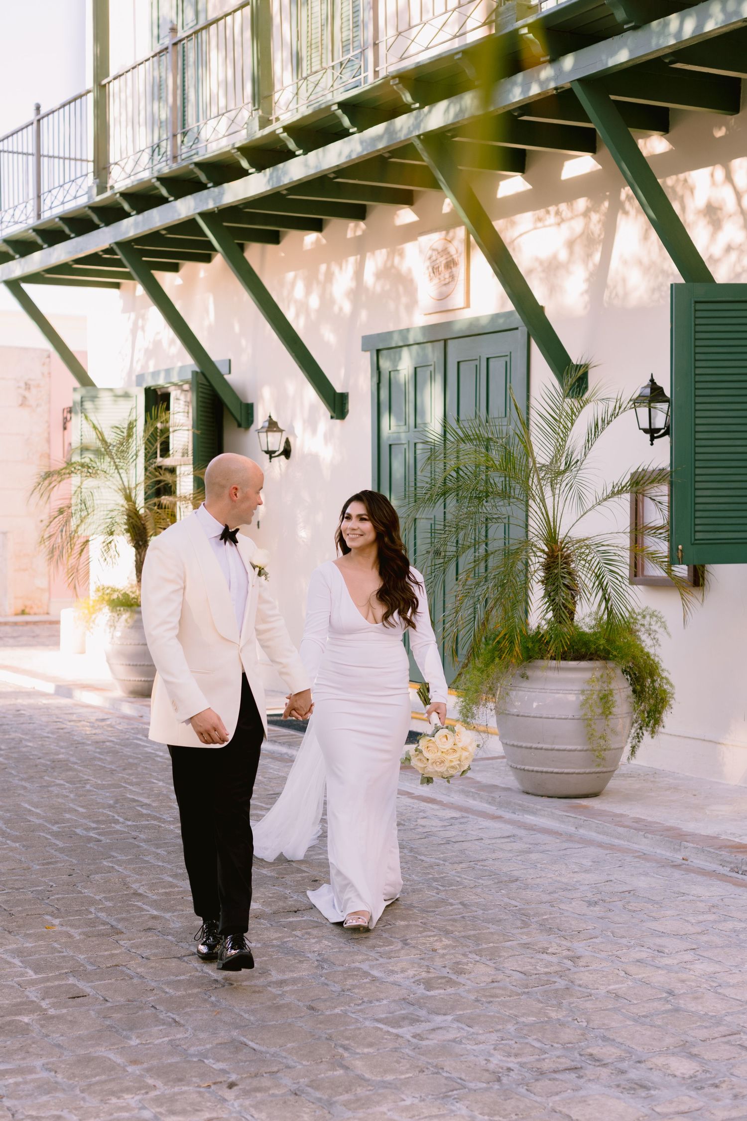 Two people in white formal attire walk together on a stone courtyard with white walls and green shutters in the background.