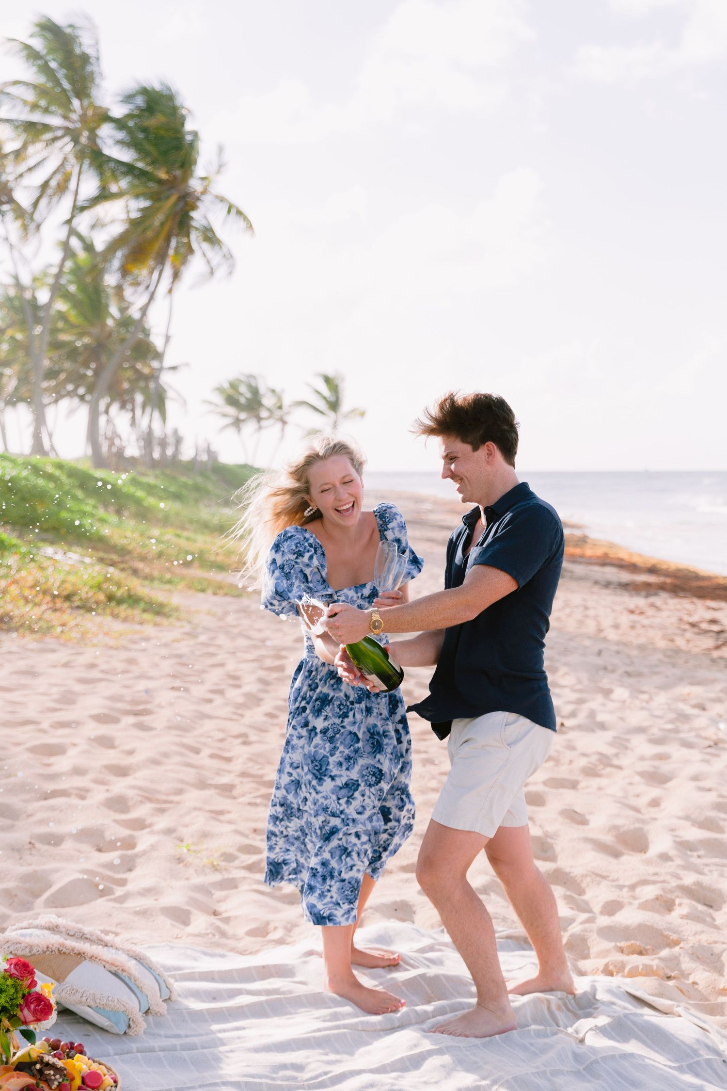 Two people playfully dance together on a tropical beach with palm trees swaying in the wind.