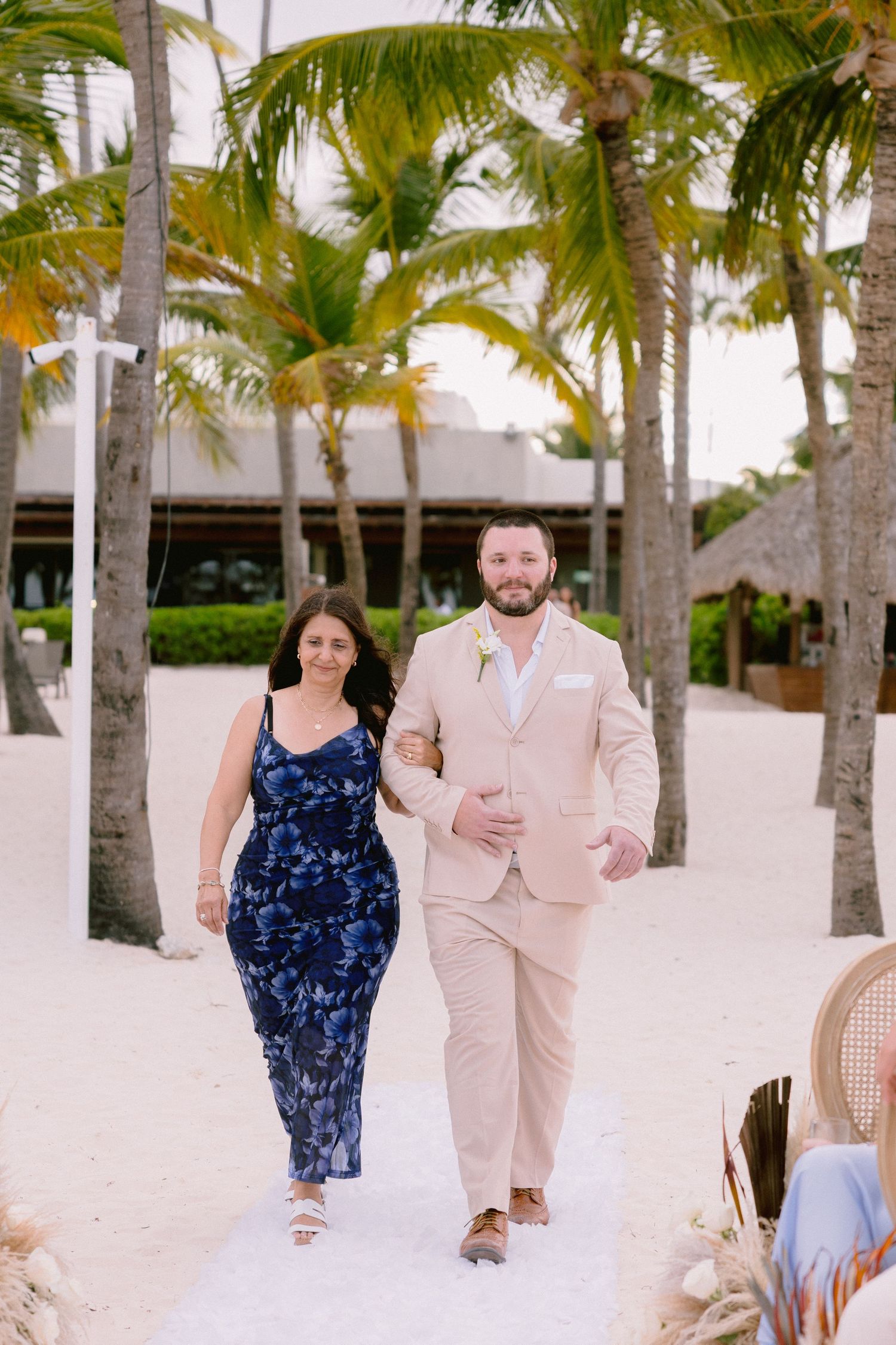 Couple walking down beach aisle, man in tan suit with boutonniere, woman in navy floral dress, palm trees backdrop.