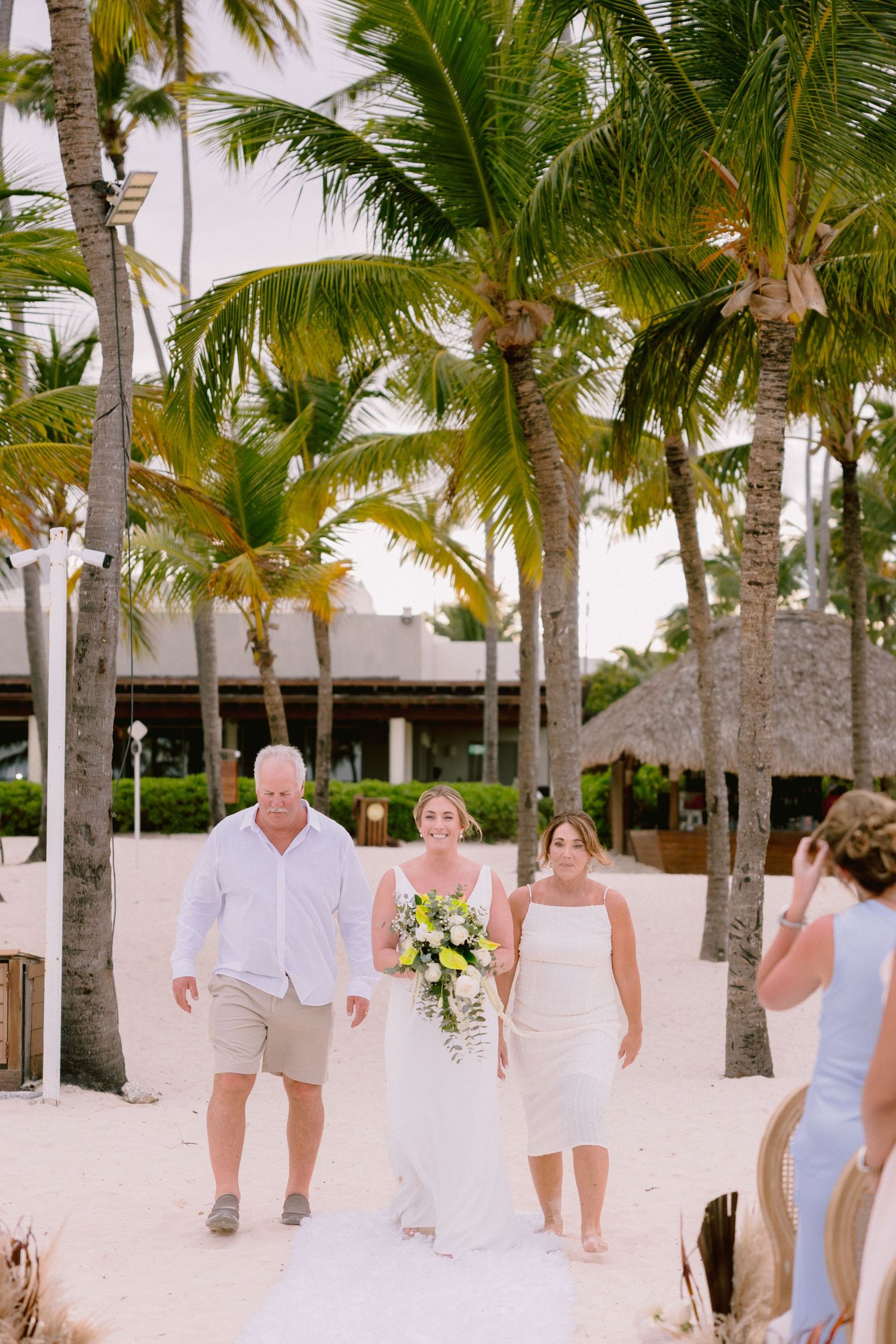 Bride walks down sandy beach aisle with family, surrounded by palm trees at a tropical destination wedding.