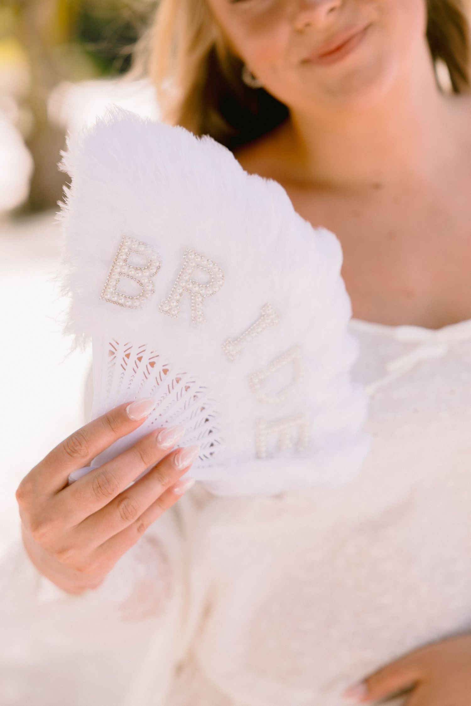 Bride holding a white feathered fan with pearl letters spelling BRIDE at her wedding celebration.