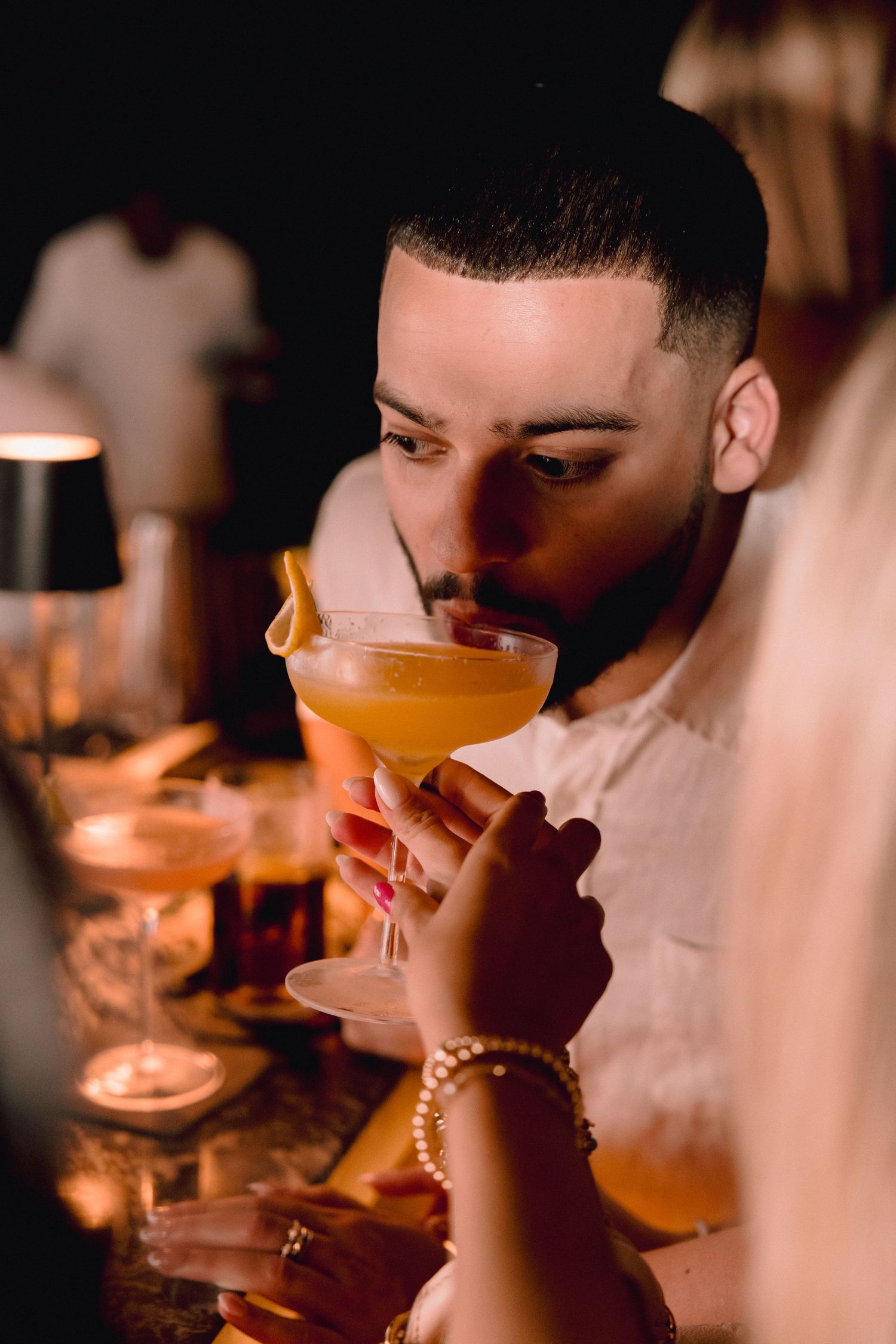 A man in a white shirt sips an orange cocktail from a coupe glass at a dimly lit bar while a woman with bracelets sits beside him.