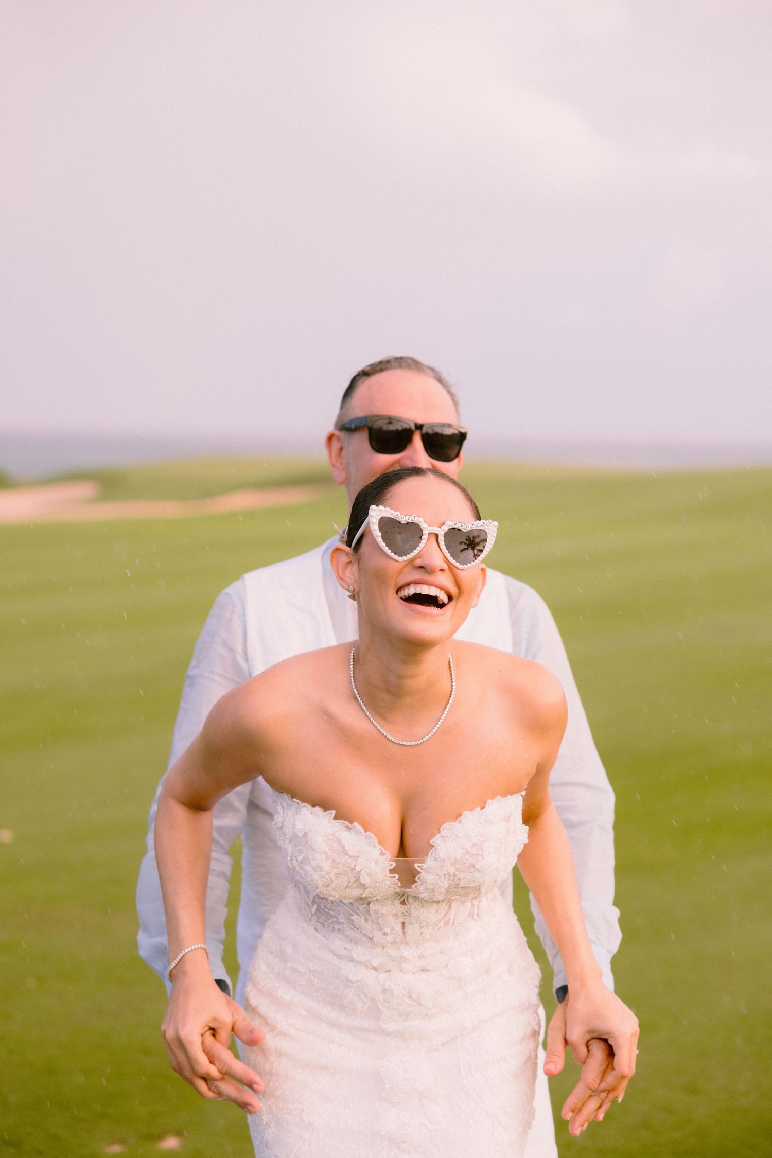 A joyful bride wearing sunglasses and a white wedding dress runs through a green field on a sunny day.