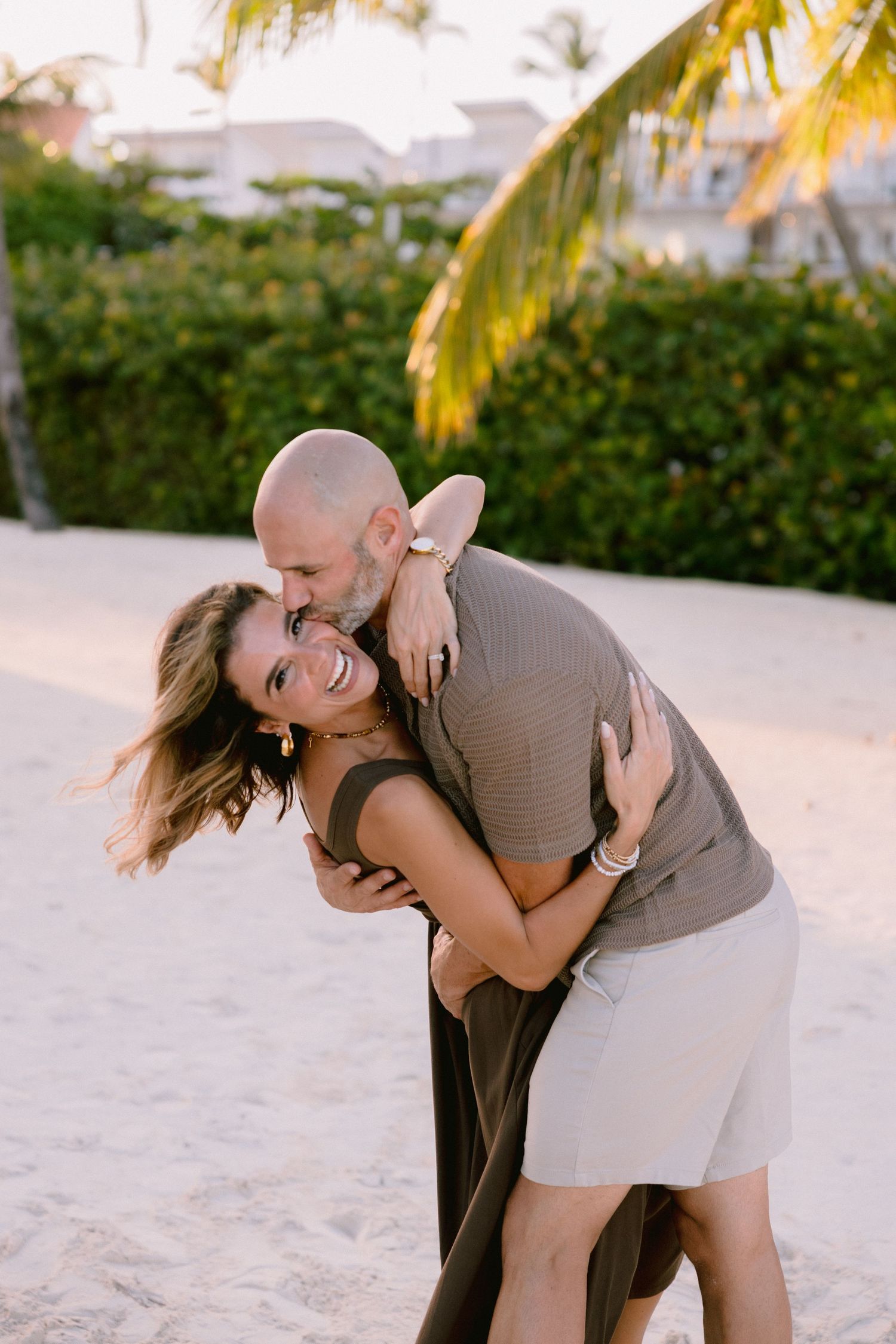 A couple embraces playfully on a tropical beach with palm trees and buildings in the background during sunset.