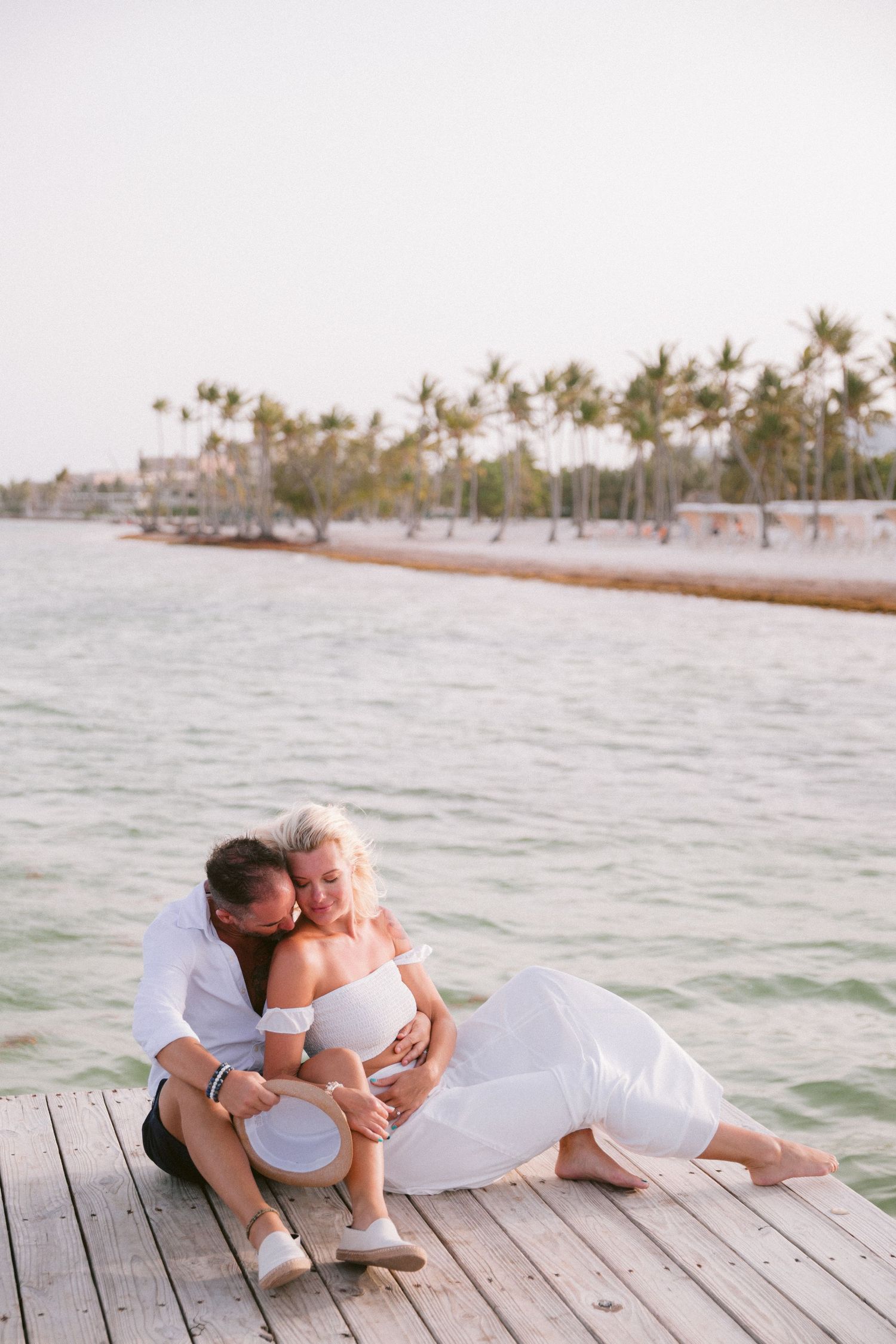 A couple in white clothing lying together on a wooden dock by the ocean at sunset.