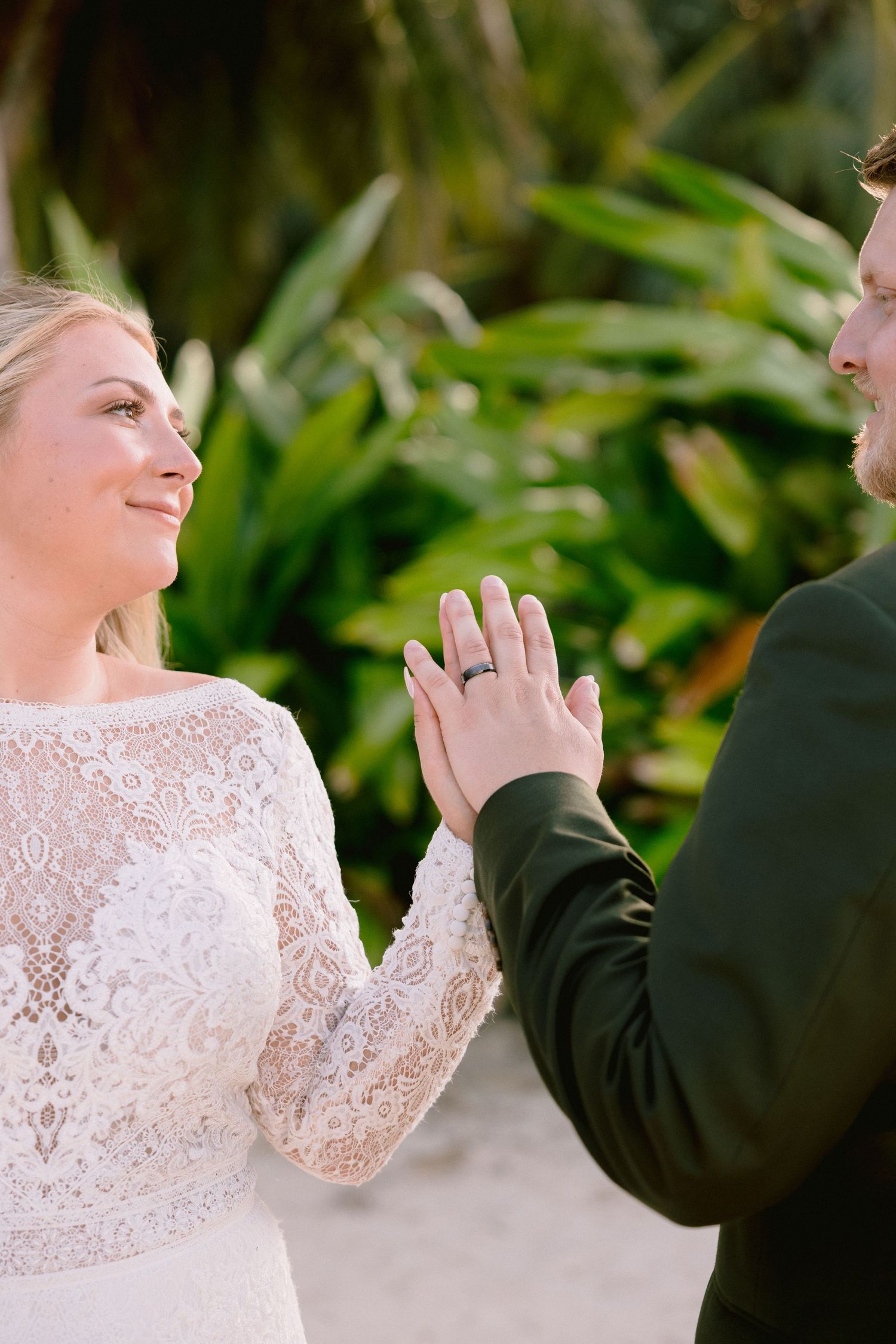 Bride in lace gown and groom in dark suit exchange rings outdoors with tropical greenery backdrop.