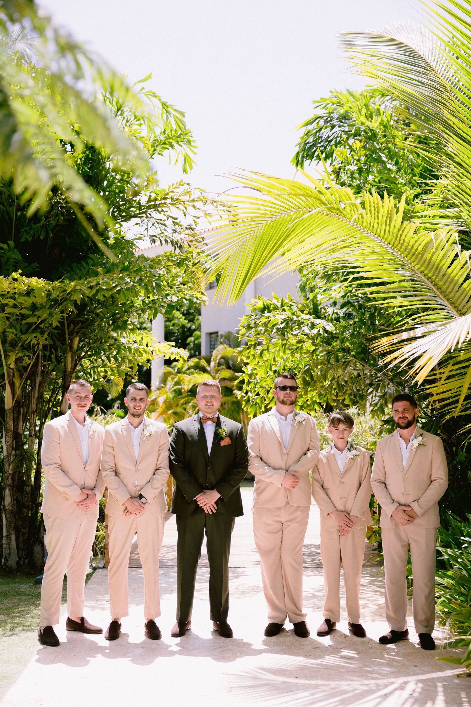 Groom in olive suit with groomsmen in tan suits posing outdoors among lush tropical greenery at a destination wedding.