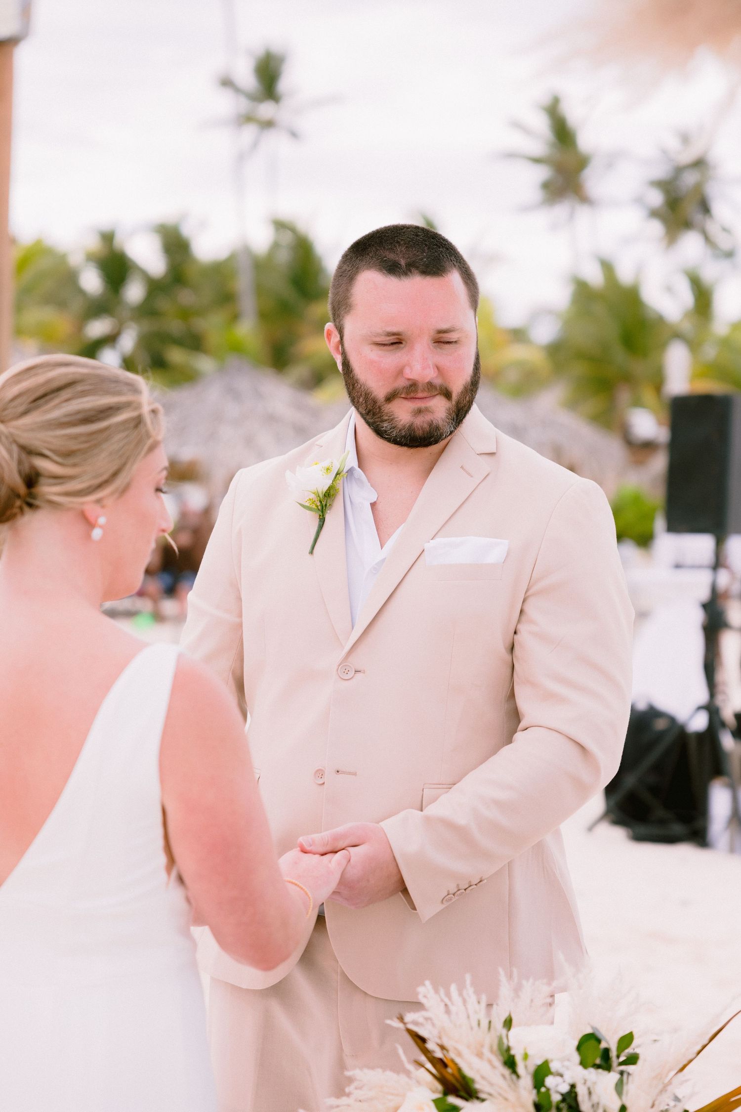 Groom in beige suit holds bride's hands during outdoor beach wedding ceremony with tropical palm trees.