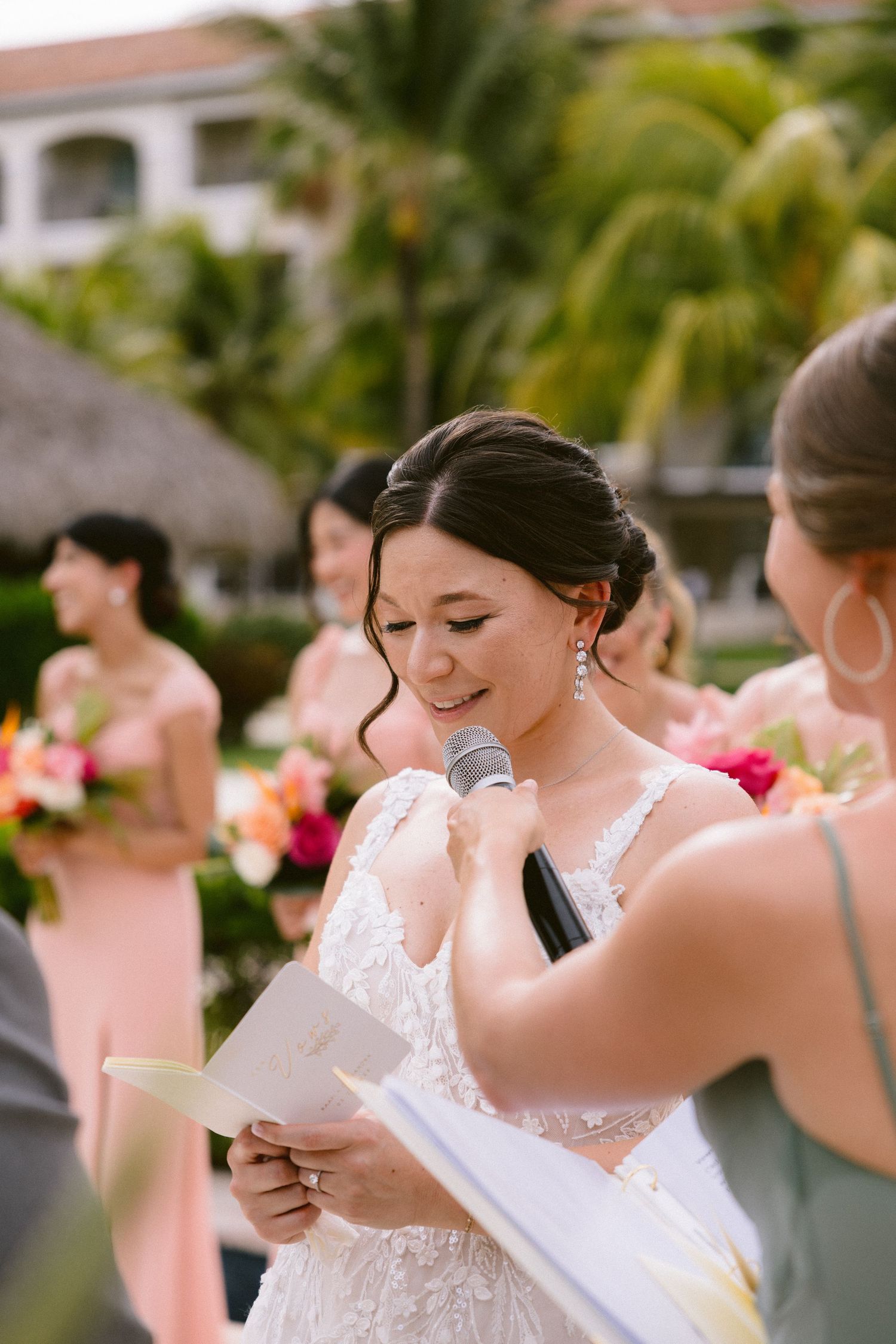 Bride in white lace wedding dress reads from paper during outdoor ceremony with pink-dressed bridesmaids and palm trees.