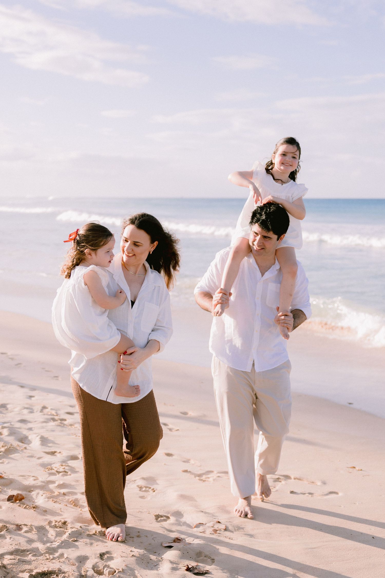 Happy family of four walking barefoot on a sunny beach, two adults in white shirts carrying children on a sandy shore.