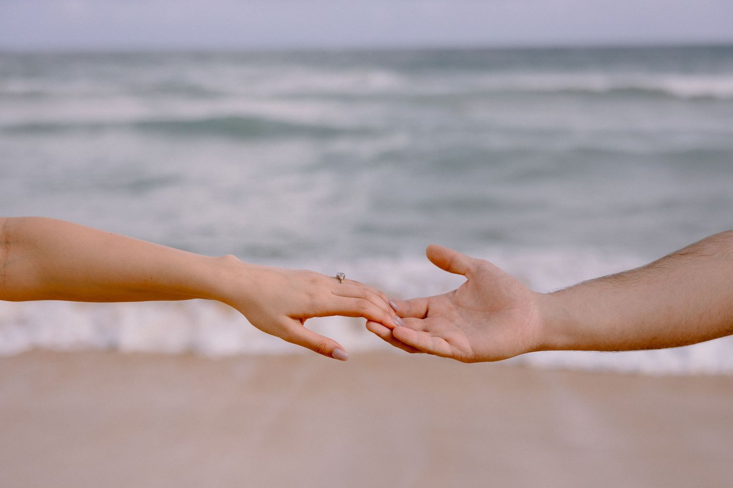 Two hands gently touching fingertips on a beach with ocean waves in the background.