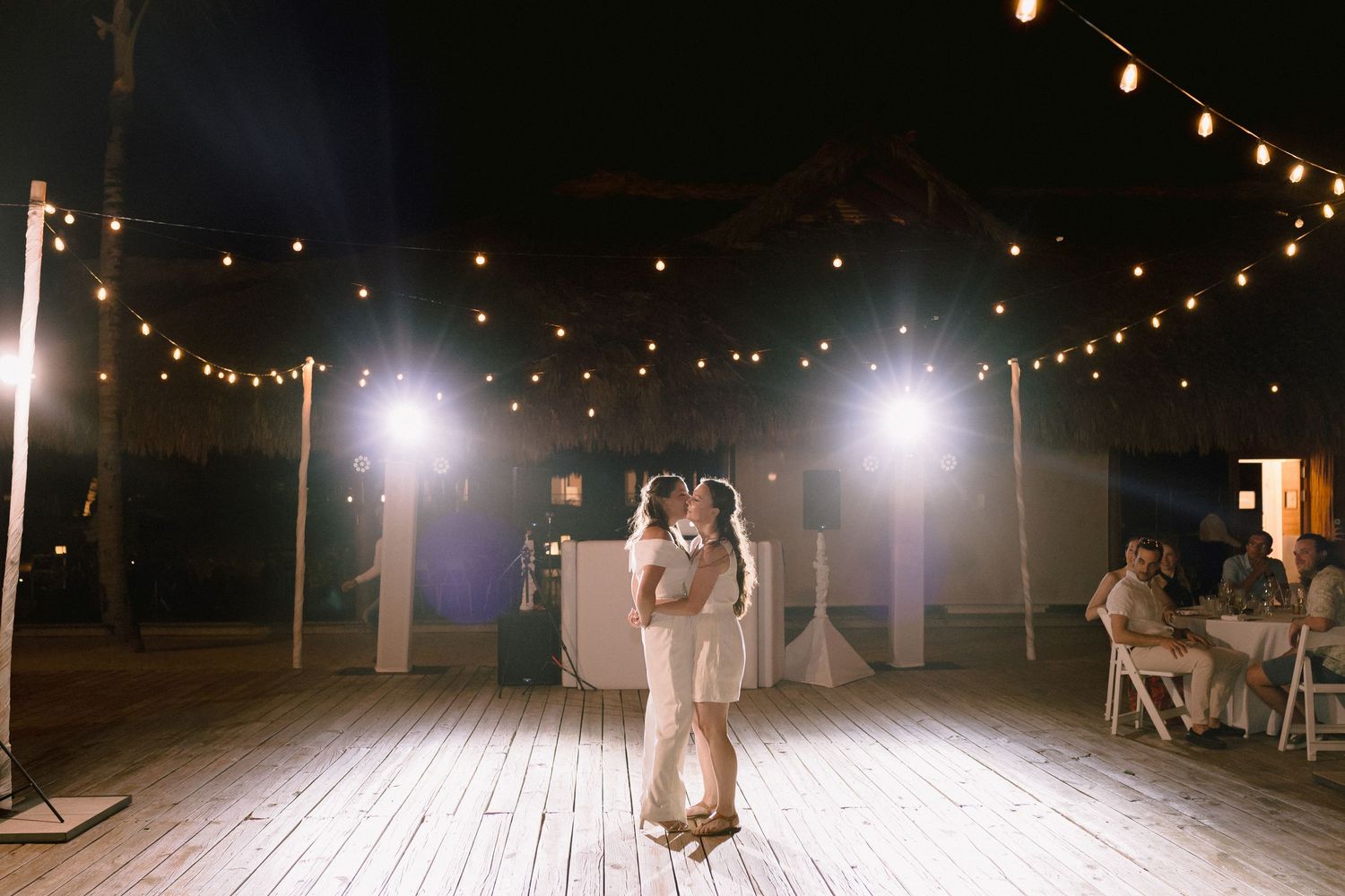 Couple shares first dance on wooden floor under string lights at outdoor wedding reception at night.
