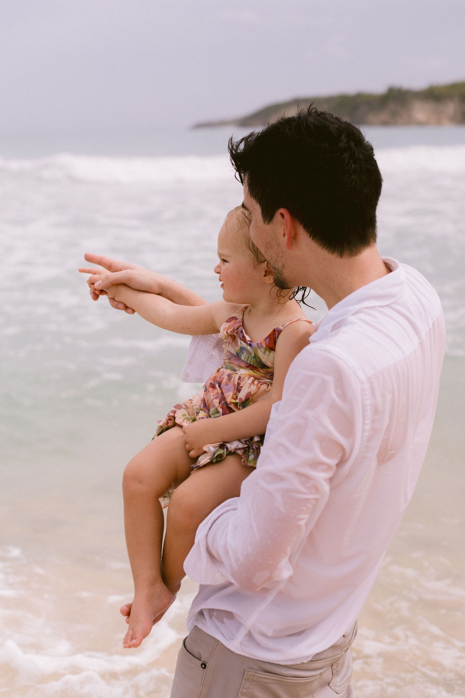 Father holding young daughter on a beach, as she points toward the ocean waves on a cloudy day.